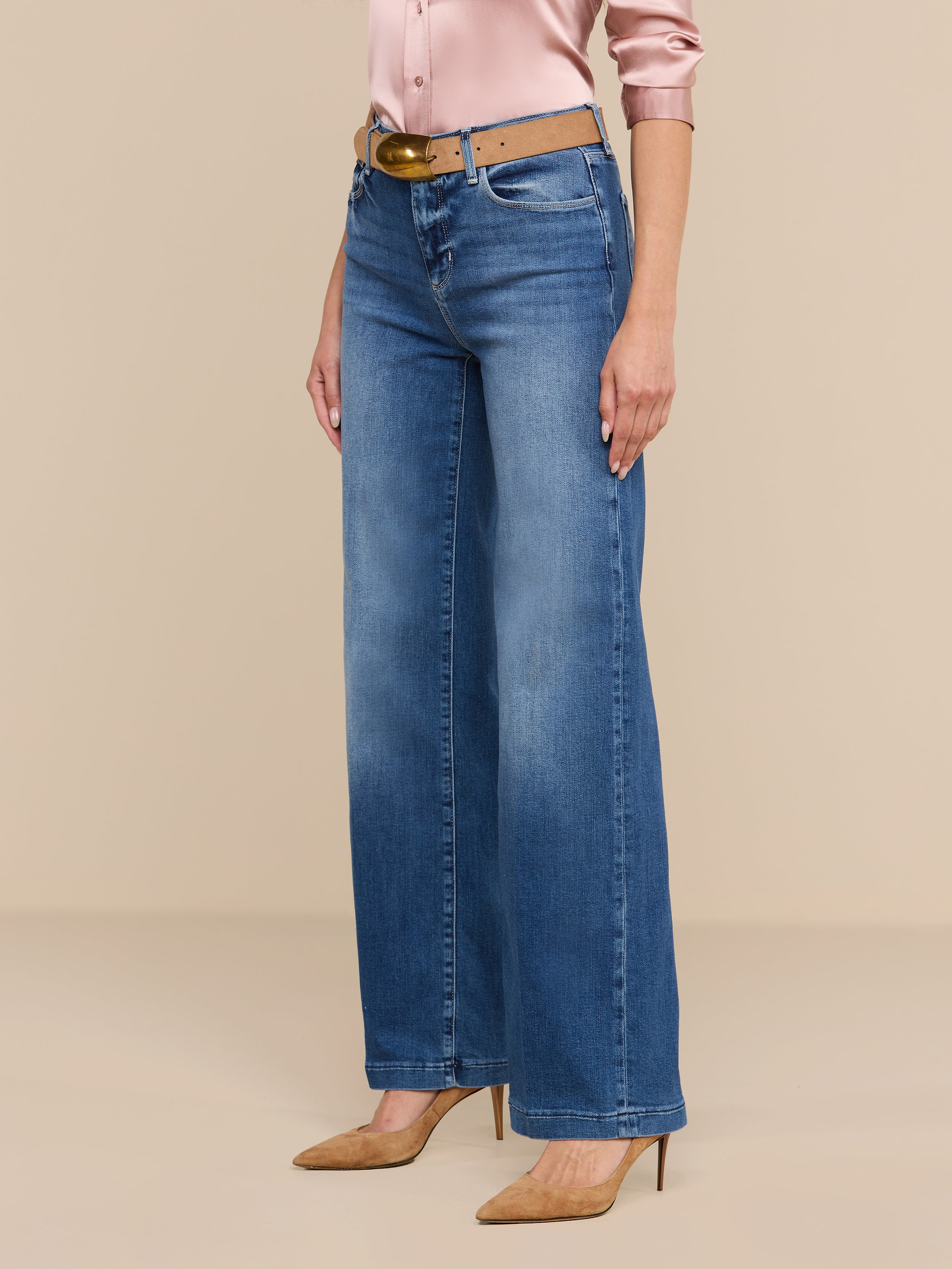 Donna che indossa i jeans Scottie Wide-Leg in denim elasticizzato blu, con una cintura marrone, una camicetta di raso rosa chiaro e scarpe con tacco a punta color cuoio, in piedi su uno sfondo beige.