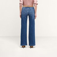 Si vede una persona di spalle che indossa un paio di jeans Scottie Wide-Leg in denim elasticizzato blu, abbinati a una camicetta a maniche lunghe rosa chiaro e a scarpe con tacco marroni, in piedi su uno sfondo neutro.