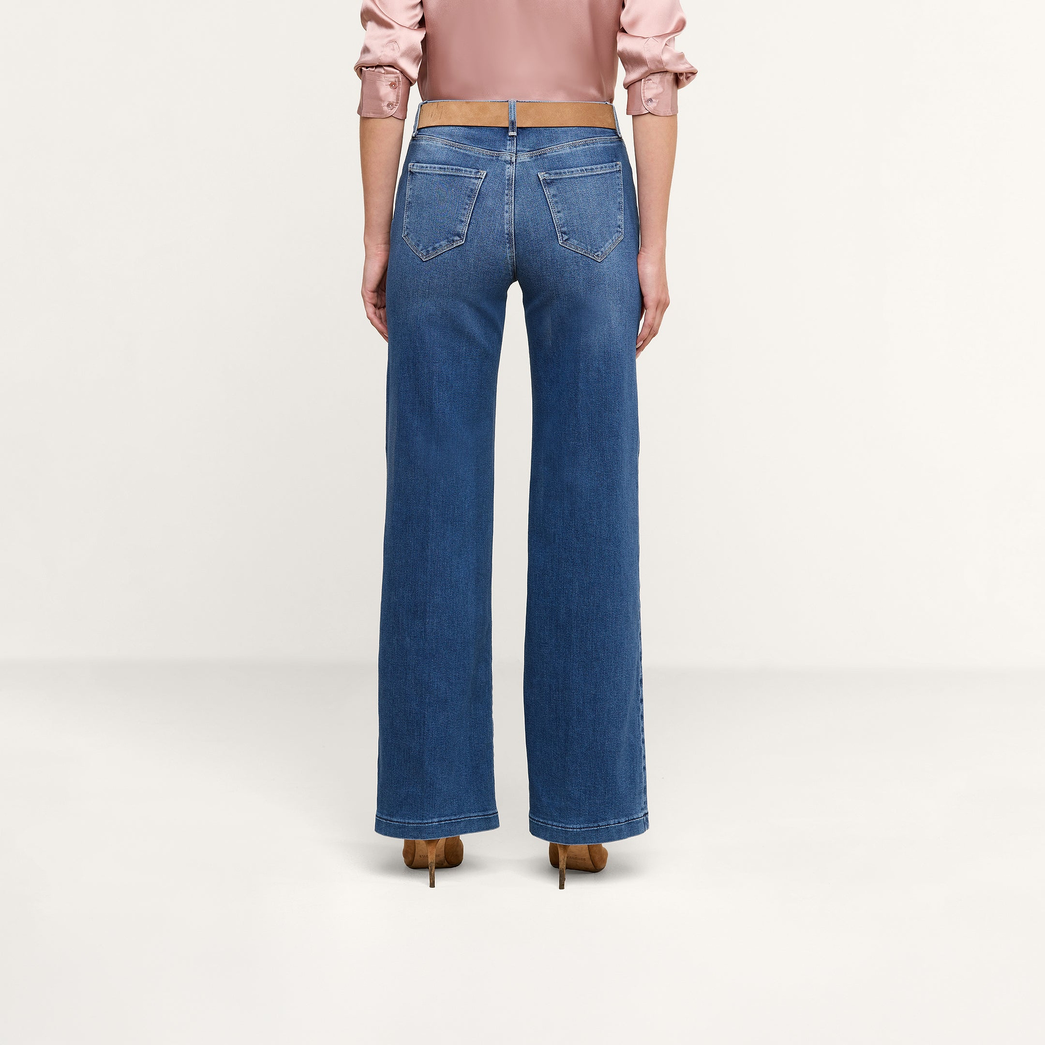 Si vede una persona di spalle che indossa un paio di jeans Scottie Wide-Leg in denim elasticizzato blu, abbinati a una camicetta a maniche lunghe rosa chiaro e a scarpe con tacco marroni, in piedi su uno sfondo neutro.