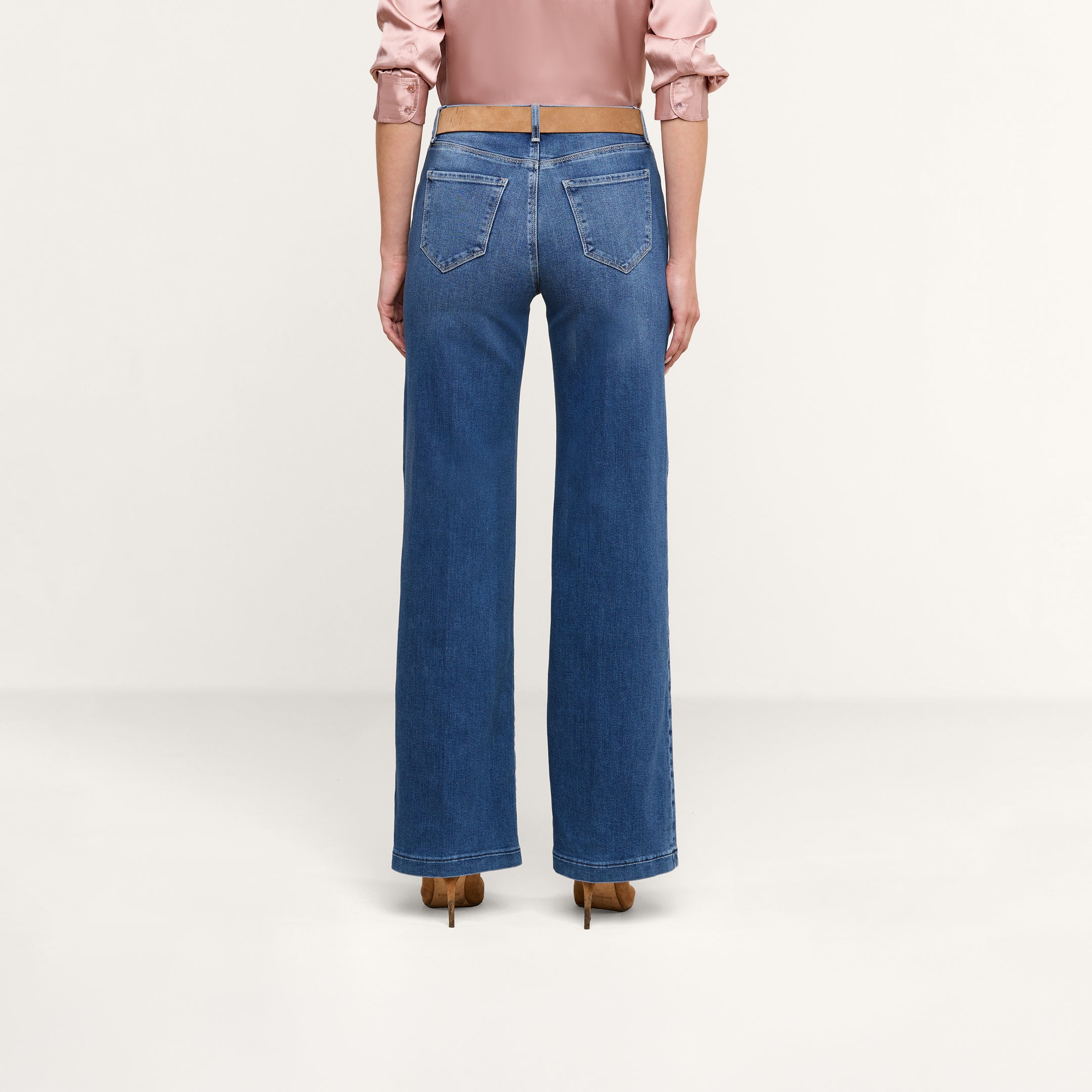 Si vede una persona di spalle che indossa un paio di jeans Scottie Wide-Leg in denim elasticizzato blu, abbinati a una camicetta a maniche lunghe rosa chiaro e a scarpe con tacco marroni, in piedi su uno sfondo neutro.