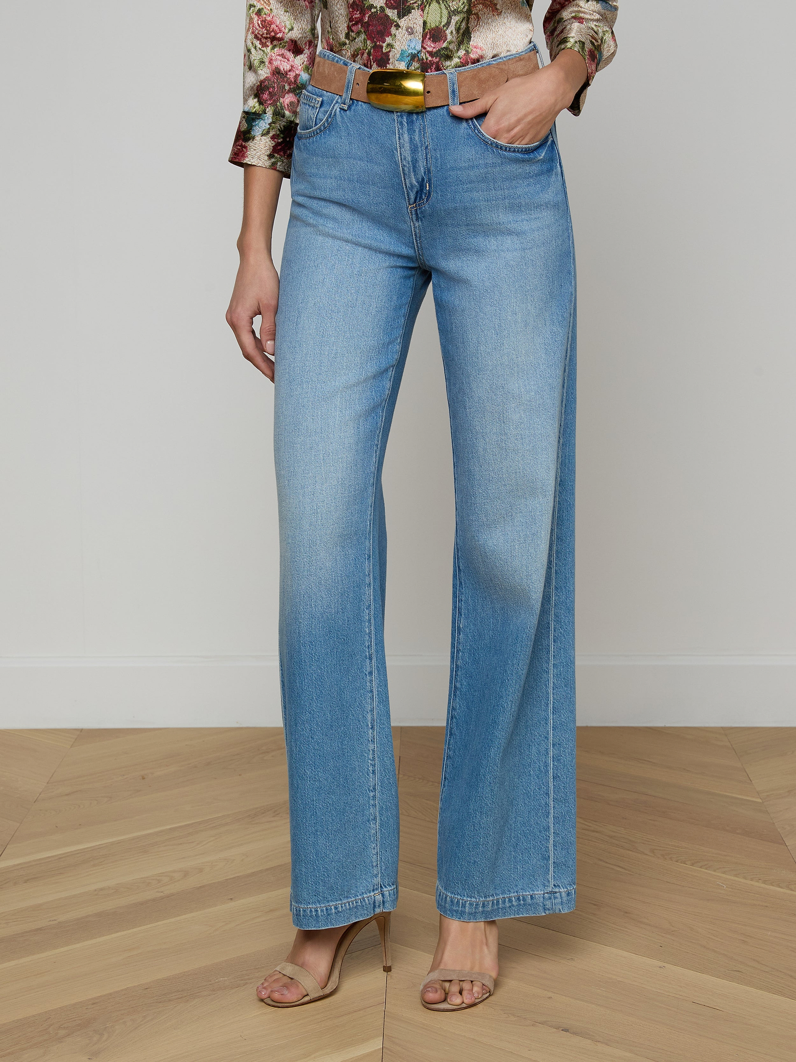Wearing the Scottie Wide-Leg Jean—high-waisted with a light wash and contoured waistband—paired with a floral long-sleeve top and tan heeled sandals, this person stands against a plain background.