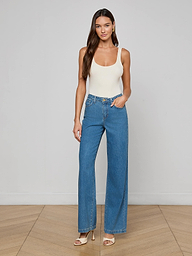 A woman stands against a plain background, wearing a white tank top, the Scottie Wide-Leg Jean in medium wash blue high-waisted all-cotton denim, and beige heeled sandals. She has long hair and is posing with one hand in her pocket.