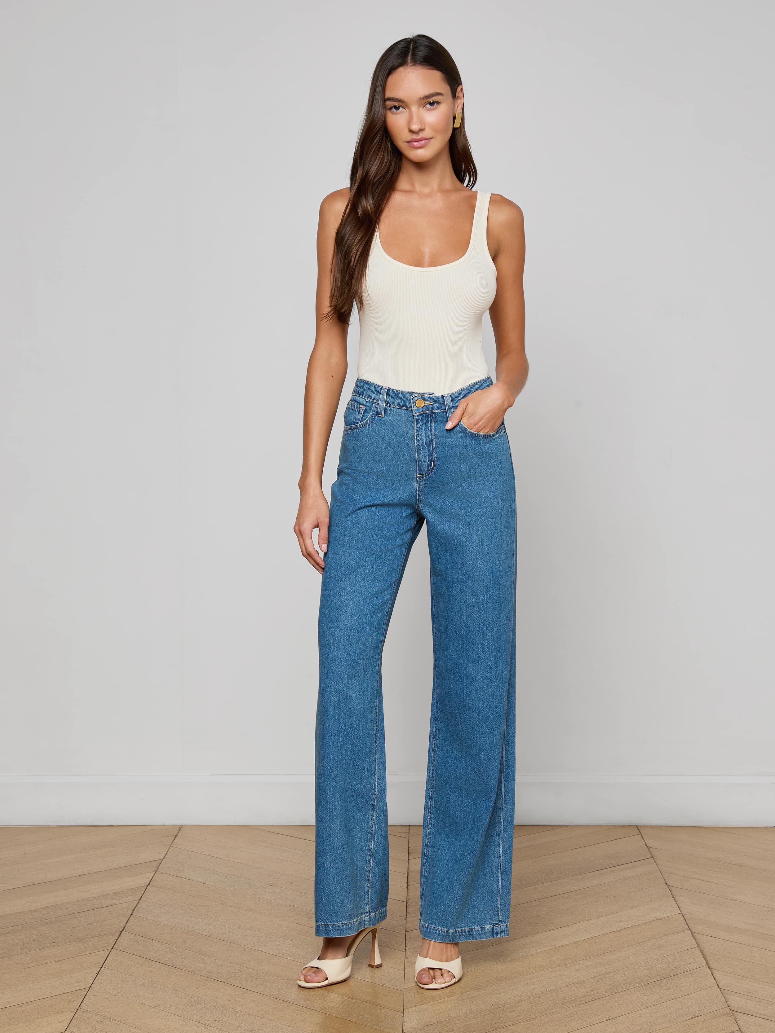 A woman stands against a plain background, wearing a white tank top, the Scottie Wide-Leg Jean in medium wash blue high-waisted all-cotton denim, and beige heeled sandals. She has long hair and is posing with one hand in her pocket.