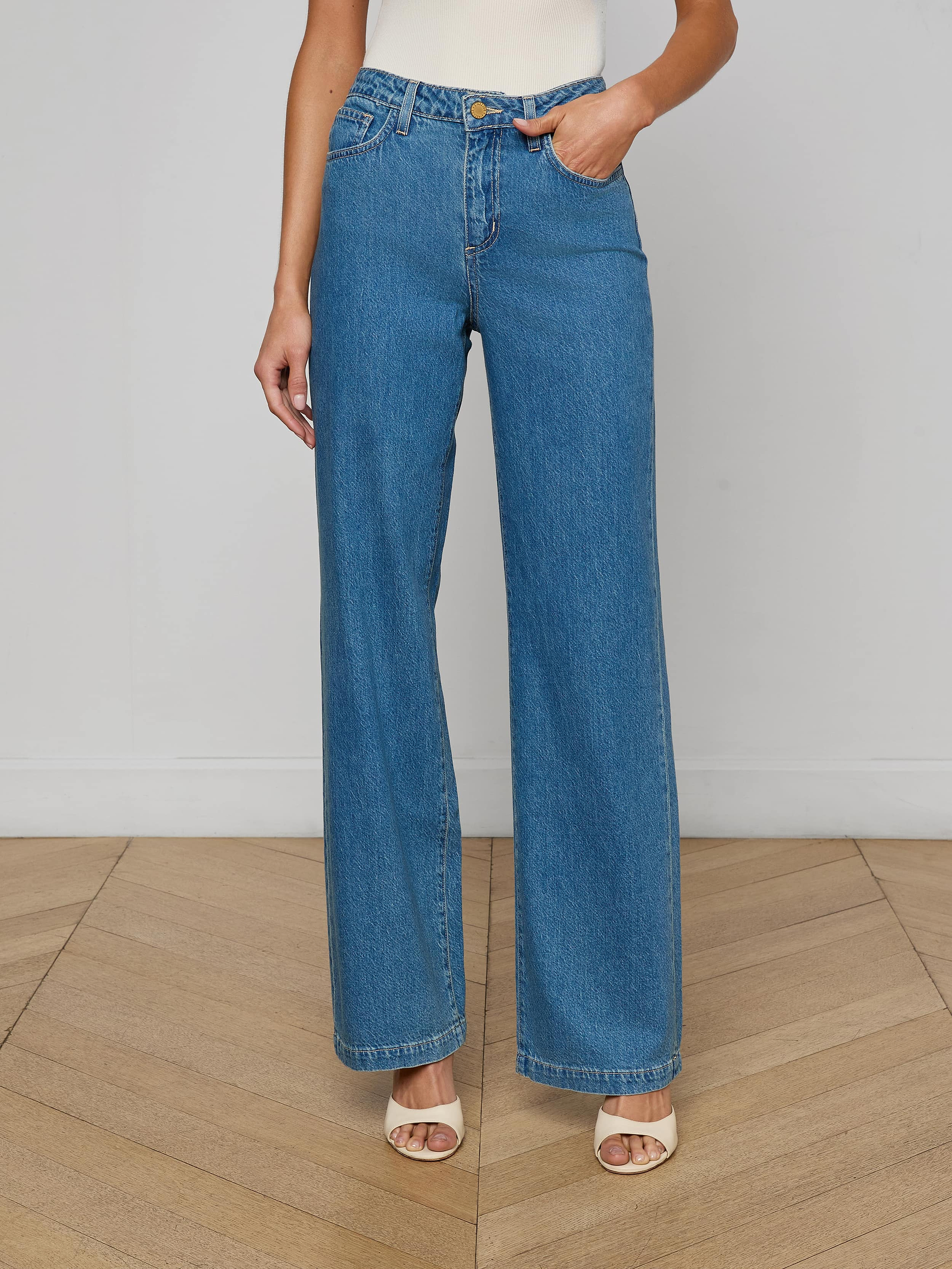 A person shown from the shoulders down against a plain light background wears Scottie Wide-Leg Jean in medium wash, crafted from all-cotton denim, paired with open-toe beige heels.