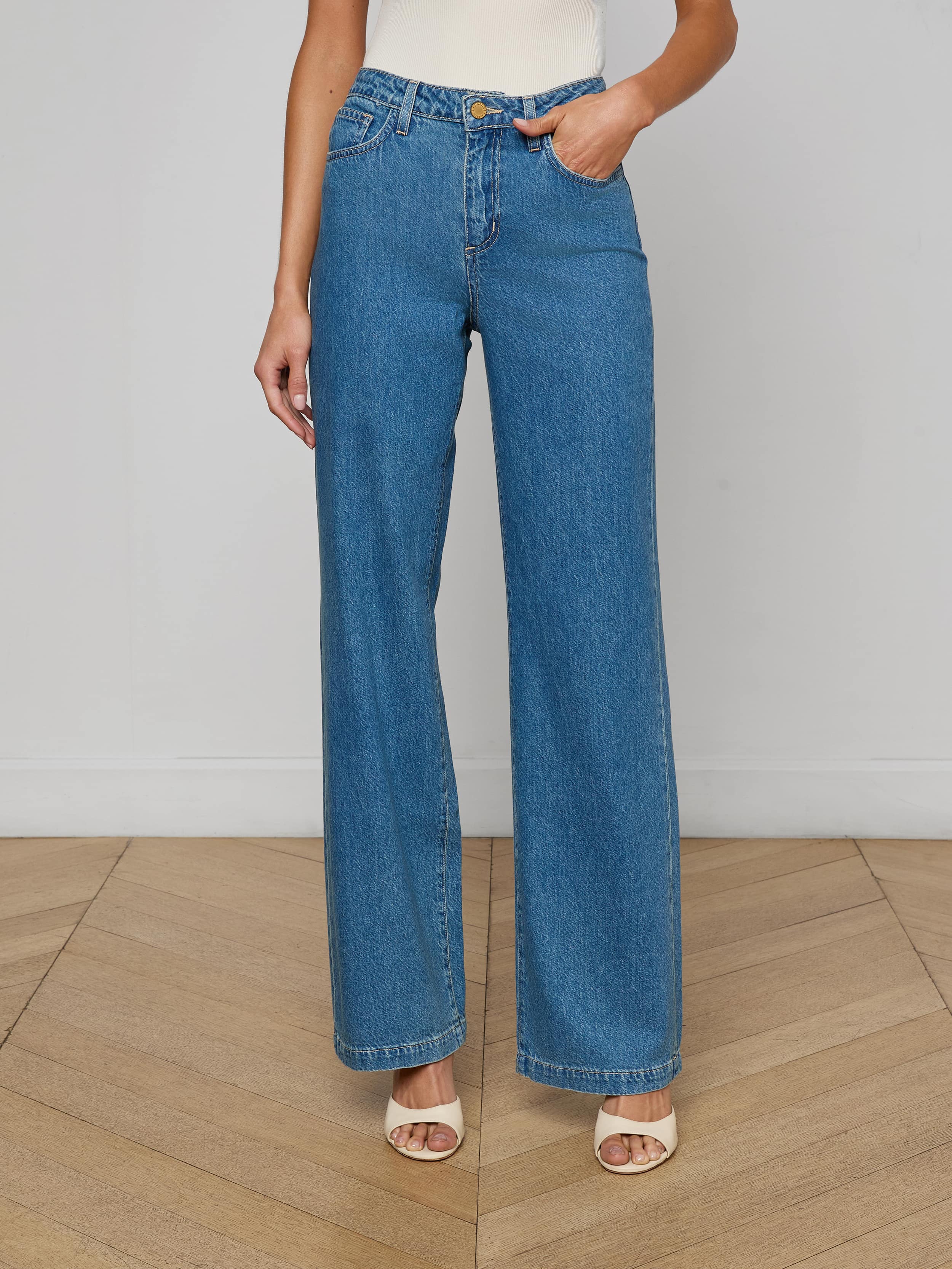 A person shown from the shoulders down against a plain light background wears Scottie Wide-Leg Jean in medium wash, crafted from all-cotton denim, paired with open-toe beige heels.