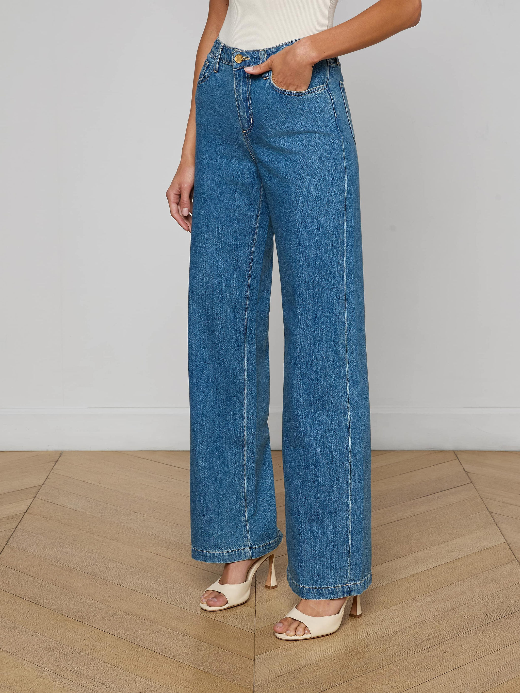 A person wearing the Scottie Wide-Leg Jean, a high-waisted, medium wash style made from 100 cotton denim, paired with open-toe beige heels stands against a plain, light background. Only the lower half of the body is visible.