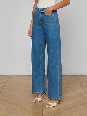 Someone stands on a wooden floor against a white wall, wearing the Scottie Wide-Leg Jean in medium wash all-cotton denim with cream heeled sandals. The top of the outfit is partly visible.