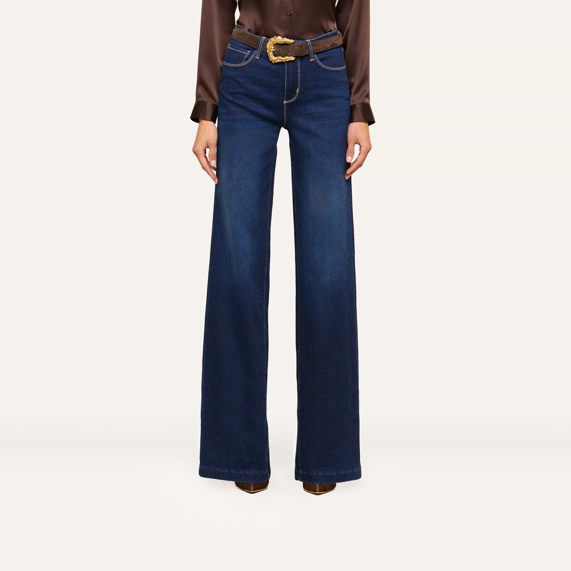 A person wearing high-waisted, wide-leg blue jeans and a brown long-sleeve blouse styled with a brown belt featuring a gold buckle and brown heeled boots stands confidently against a white background.