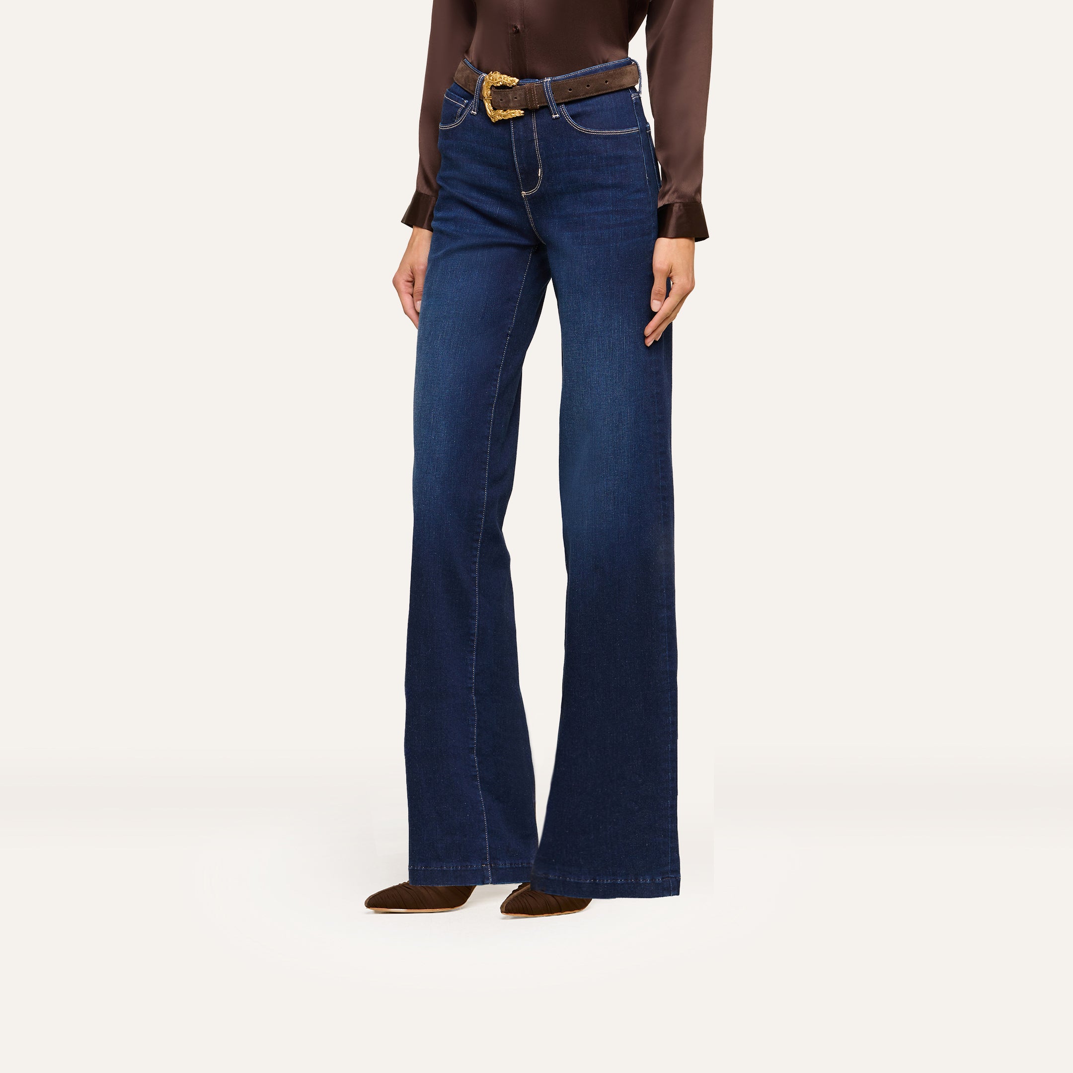 A person wearing a brown satin blouse, dark blue wide-leg jeans, a brown belt, and brown heeled boots stands against a plain white background.