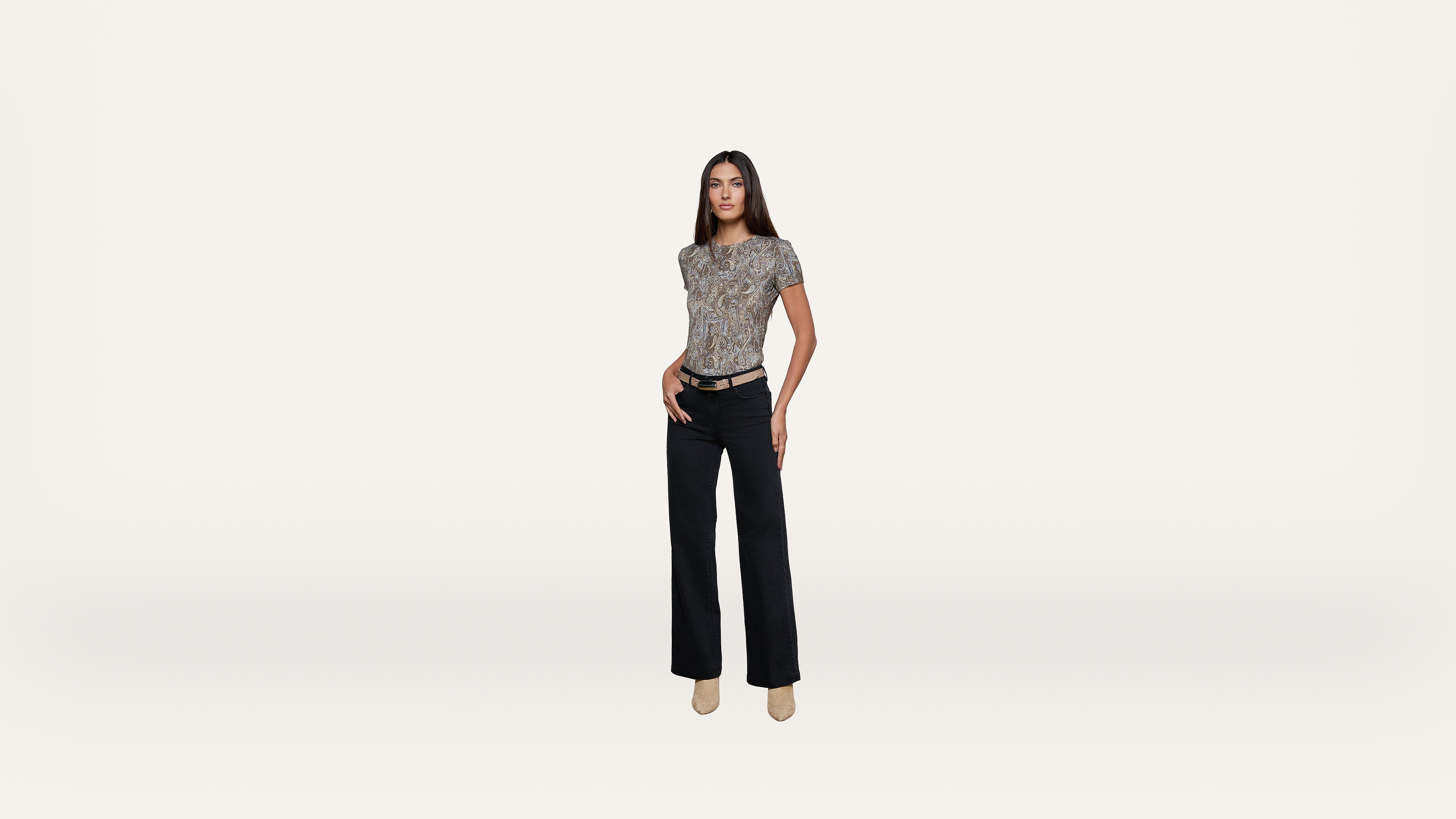 A woman stands confidently against a plain white background, wearing a patterned short-sleeve top, black pants, a belt, and beige heeled shoes.