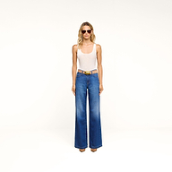 A woman stands against a plain white background wearing sunglasses, a white tank top, and the Scottie Wide-Leg Jean in an indigo wash. She completes her look with brown shoes.