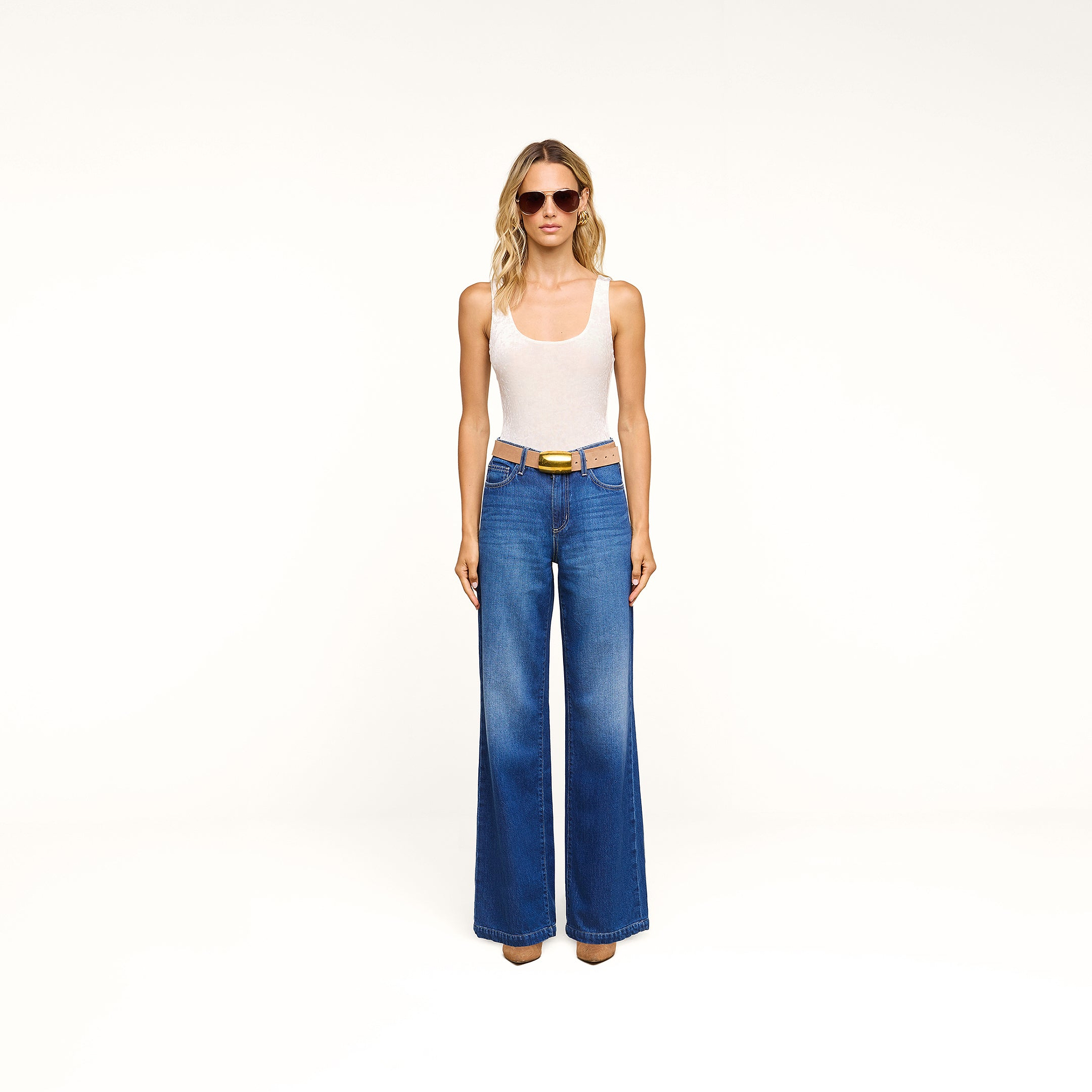 A woman stands against a plain white background wearing sunglasses, a white tank top, and the Scottie Wide-Leg Jean in an indigo wash. She completes her look with brown shoes.