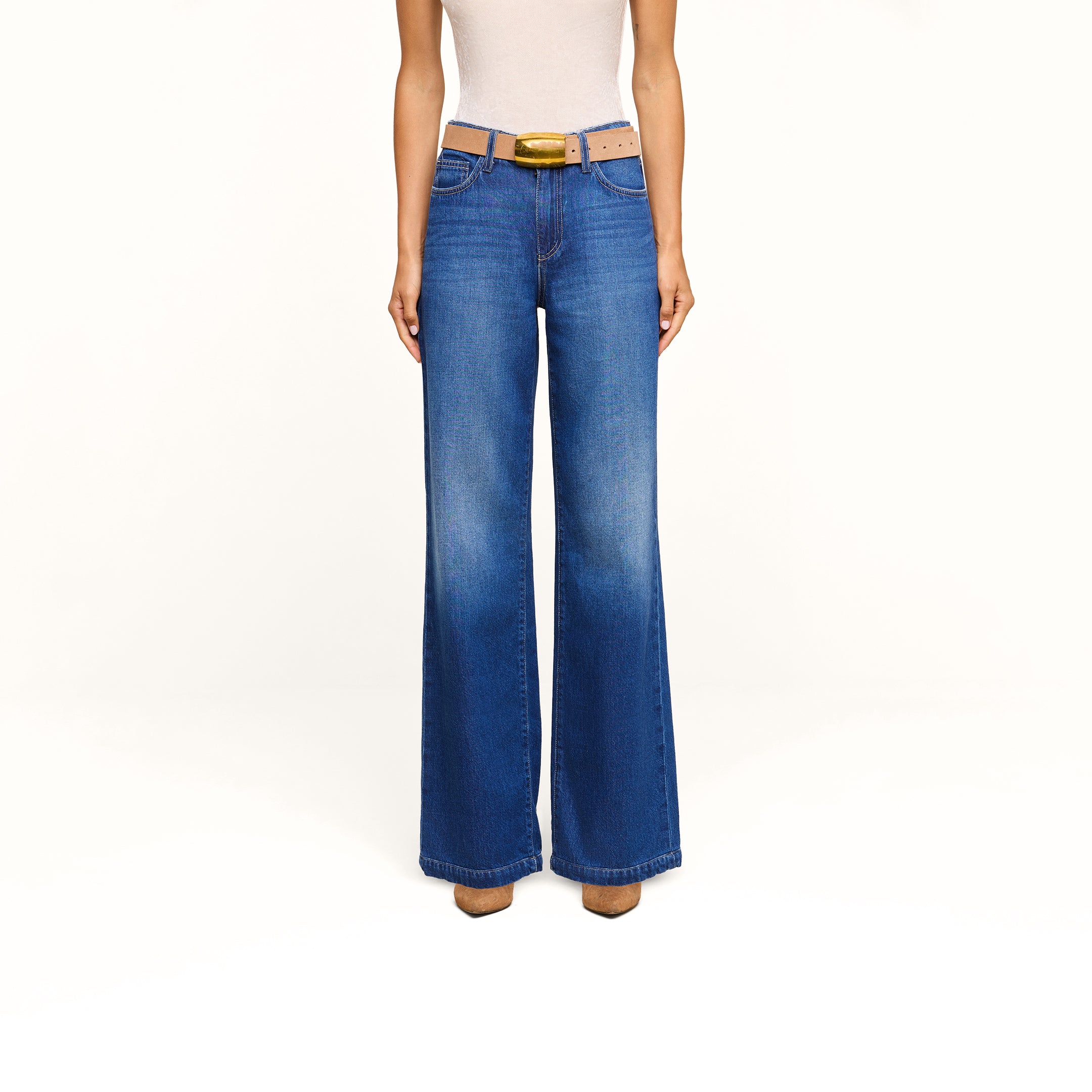 A person wearing a white sleeveless top, the Scottie Wide-Leg Jean in an indigo wash, a brown belt with a gold buckle, and brown shoes stands against a plain white background.