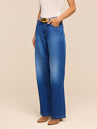 A person wearing the Scottie Wide-Leg Jean in blue nonstretch denim, paired with a white sleeveless top, a yellow belt, and tan pointed-toe shoes stands with one hand in their pocket against a beige background.