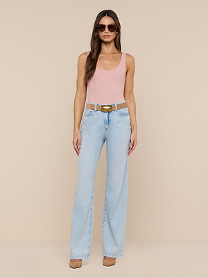 A woman wearing a light pink tank top, tan heels, sunglasses, a brown belt, and Scottie Wide-Leg Jean in a light wash stands against a plain beige background, looking straight ahead.