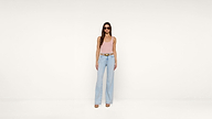 Woman wearing sunglasses, a light pink sleeveless top, light blue high-waisted wide-leg jeans, a brown belt, and beige heels stands against a plain white background.