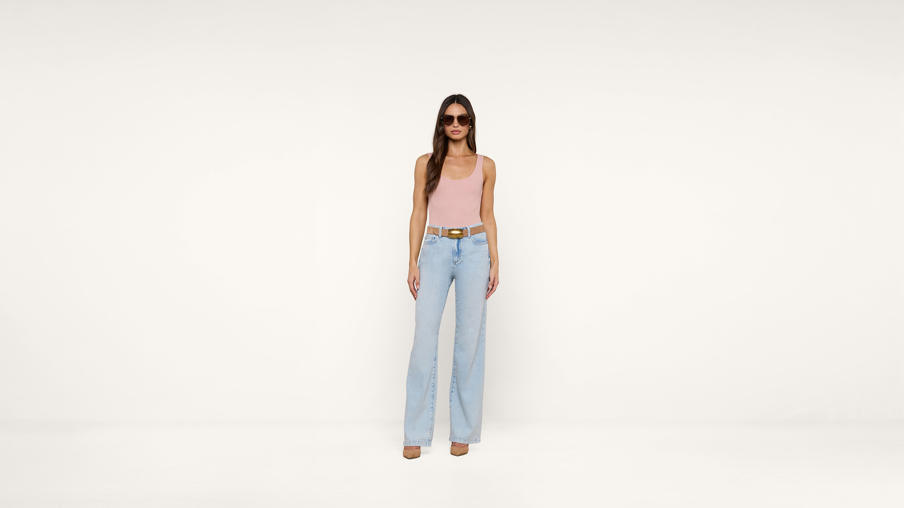 Woman wearing sunglasses, a light pink sleeveless top, light blue high-waisted wide-leg jeans, a brown belt, and beige heels stands against a plain white background.