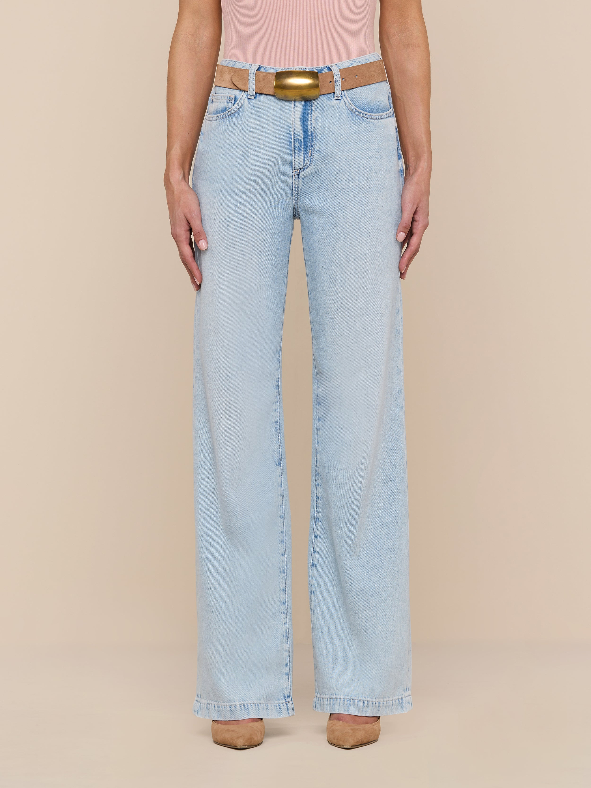 Wearing the Scottie Wide-Leg Jean in a light wash with a beige belt, a pink top, and tan shoes, the person stands against a plain white background.