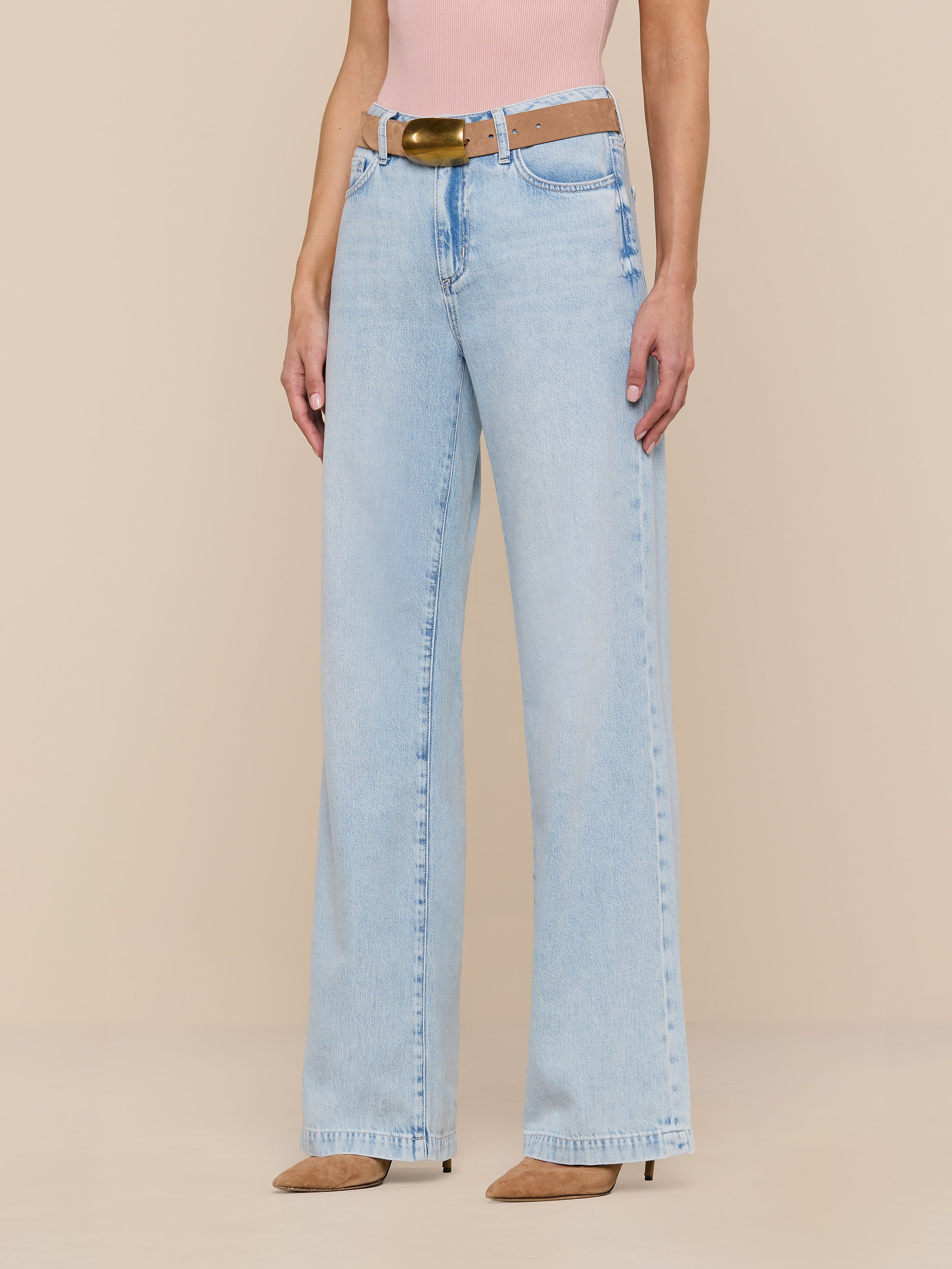 A person wearing the Scottie Wide-Leg Jean in a light wash, styled with a brown belt, a pink top, and beige pointed-toe heels stands against a plain white background.
