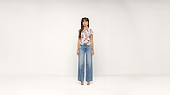 A woman stands facing forward, wearing a floral top, wide-leg jeans, a black belt, and beige sandals, against a plain white background.