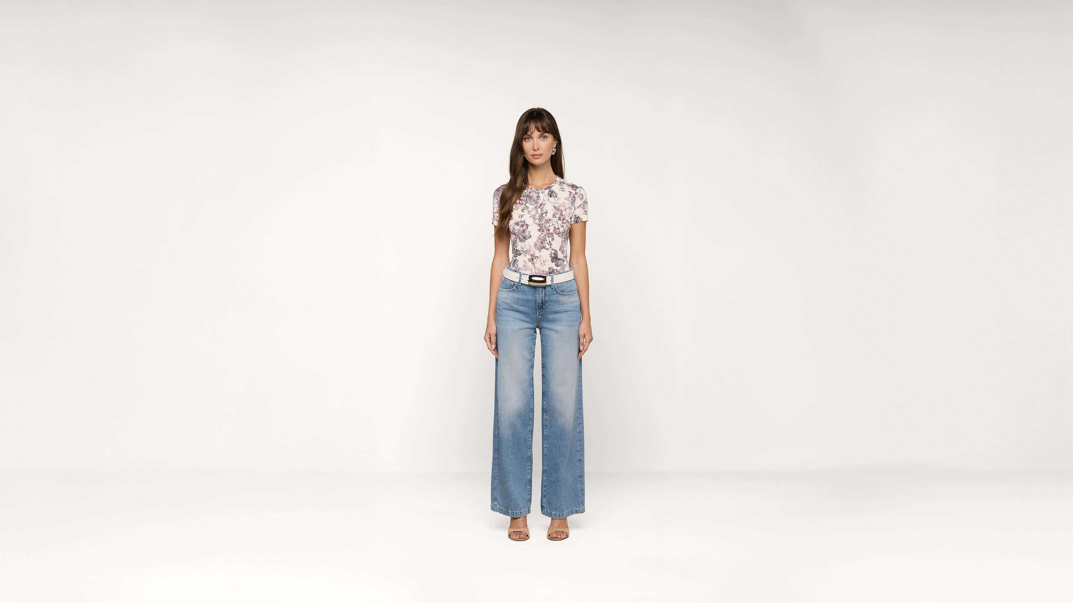 A woman stands facing forward, wearing a floral top, wide-leg jeans, a black belt, and beige sandals, against a plain white background.