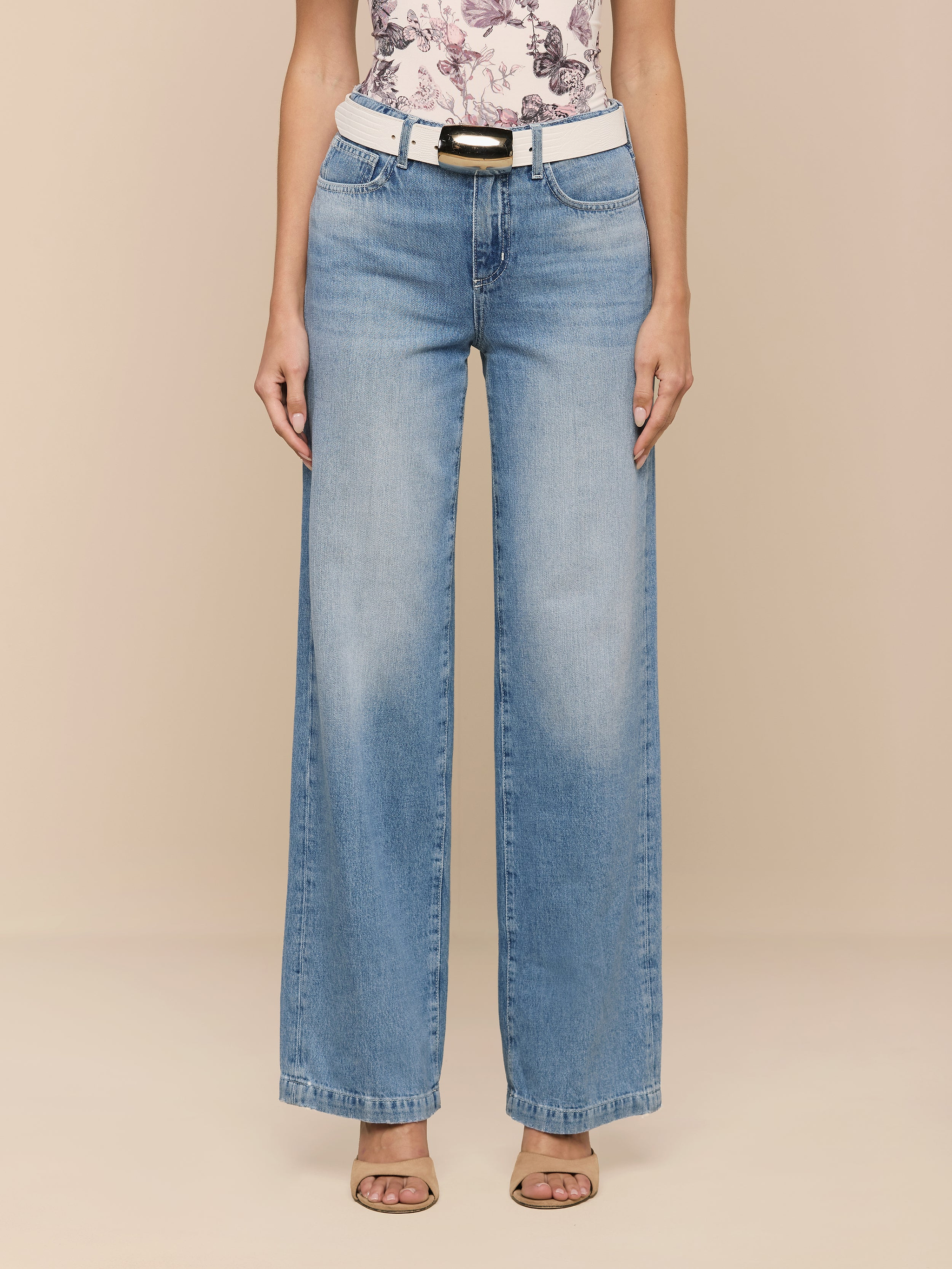Wearing the Scottie Wide-Leg Jean, a person stands against a plain white background, styled with a white belt, a floral top, and open-toe heels.