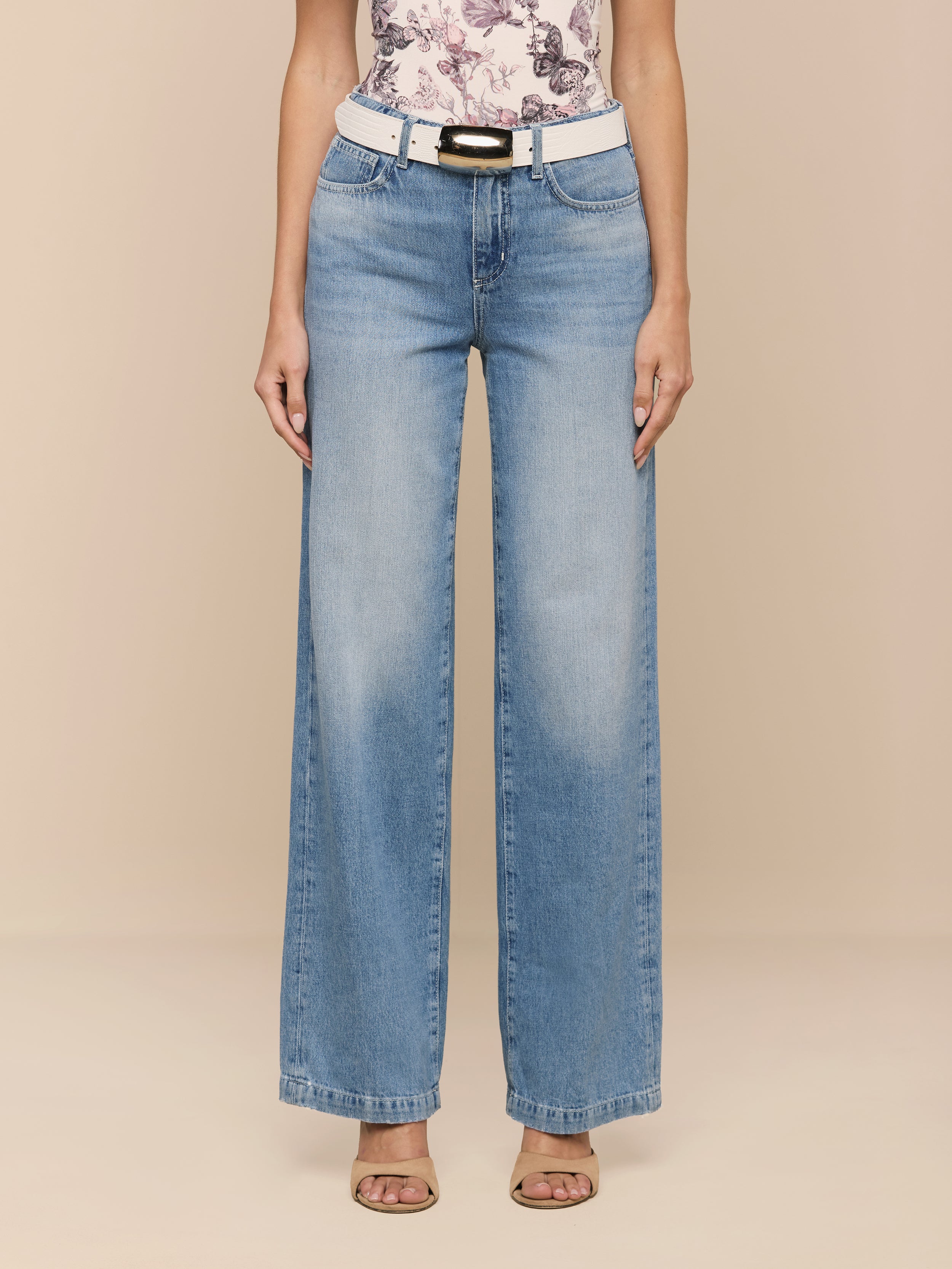 Wearing the Scottie Wide-Leg Jean, a person stands against a plain white background, styled with a white belt, a floral top, and open-toe heels.