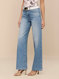 Eine Person, die Scottie Wide-Leg Jeans in hellem Indigo, einen weißen Gürtel, ein geblümtes Top und beigefarbene hochhackige Sandalen trägt, steht vor einem einfarbig beigen Hintergrund.