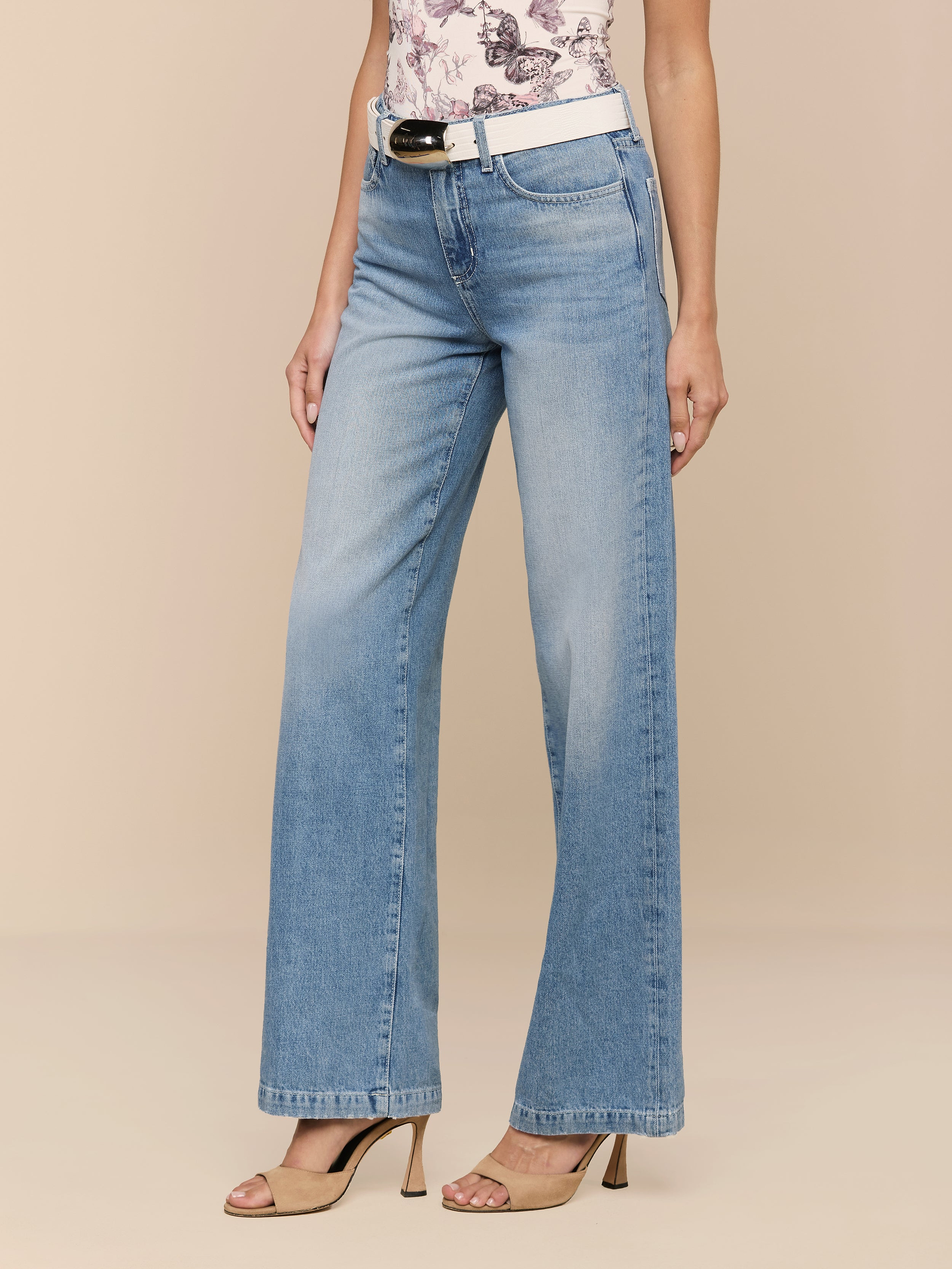 Eine Person, die Scottie Wide-Leg Jeans in hellem Indigo, einen weißen Gürtel, ein geblümtes Top und beigefarbene hochhackige Sandalen trägt, steht vor einem einfarbig beigen Hintergrund.