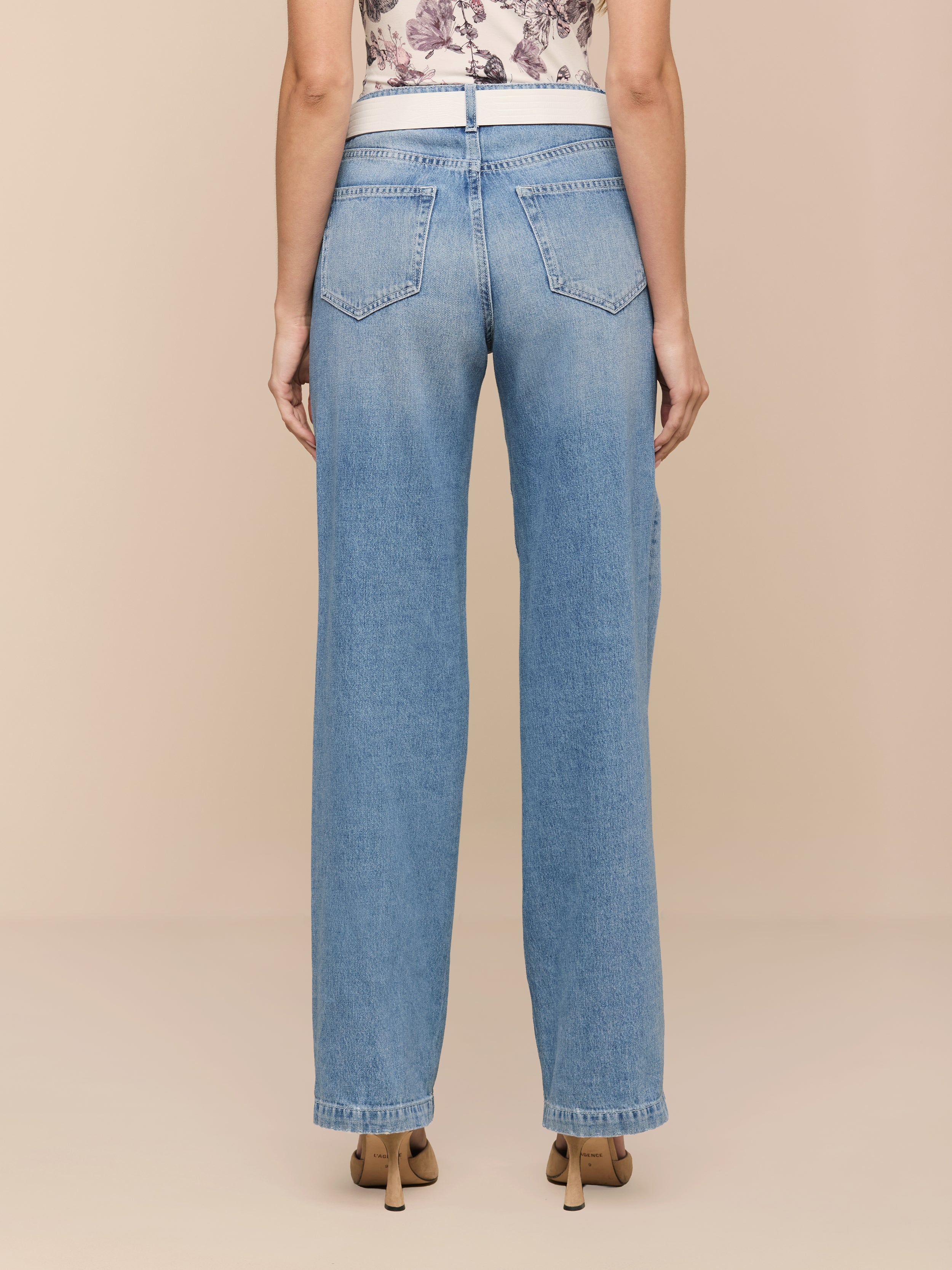 Eine Person schaut weg und trägt die Scottie Wide-Leg Jeans - eine hoch geschnittene Jeans aus reiner Baumwolle mit Gesäßtaschen und einem ärmellosen Oberteil mit Blumenmuster. Die entspannte Passform mit geradem Bein und beigefarbenen Schuhen sorgt für einen schicken, lässigen Stil.