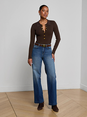 A woman with short hair and neutral makeup stands indoors against a plain wall, wearing a fitted brown long-sleeve top with gold buttons, the Scottie Wide-Leg Jean in classic stretch denim, a brown belt, and brown boots.