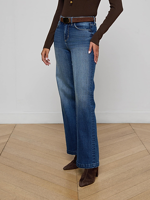 A person wearing the Scottie Wide-Leg Jean in blue denim with a contoured waistband, paired with a brown long-sleeve top, brown belt, and pointed-toe boots, stands on a wooden floor against a plain light wall.