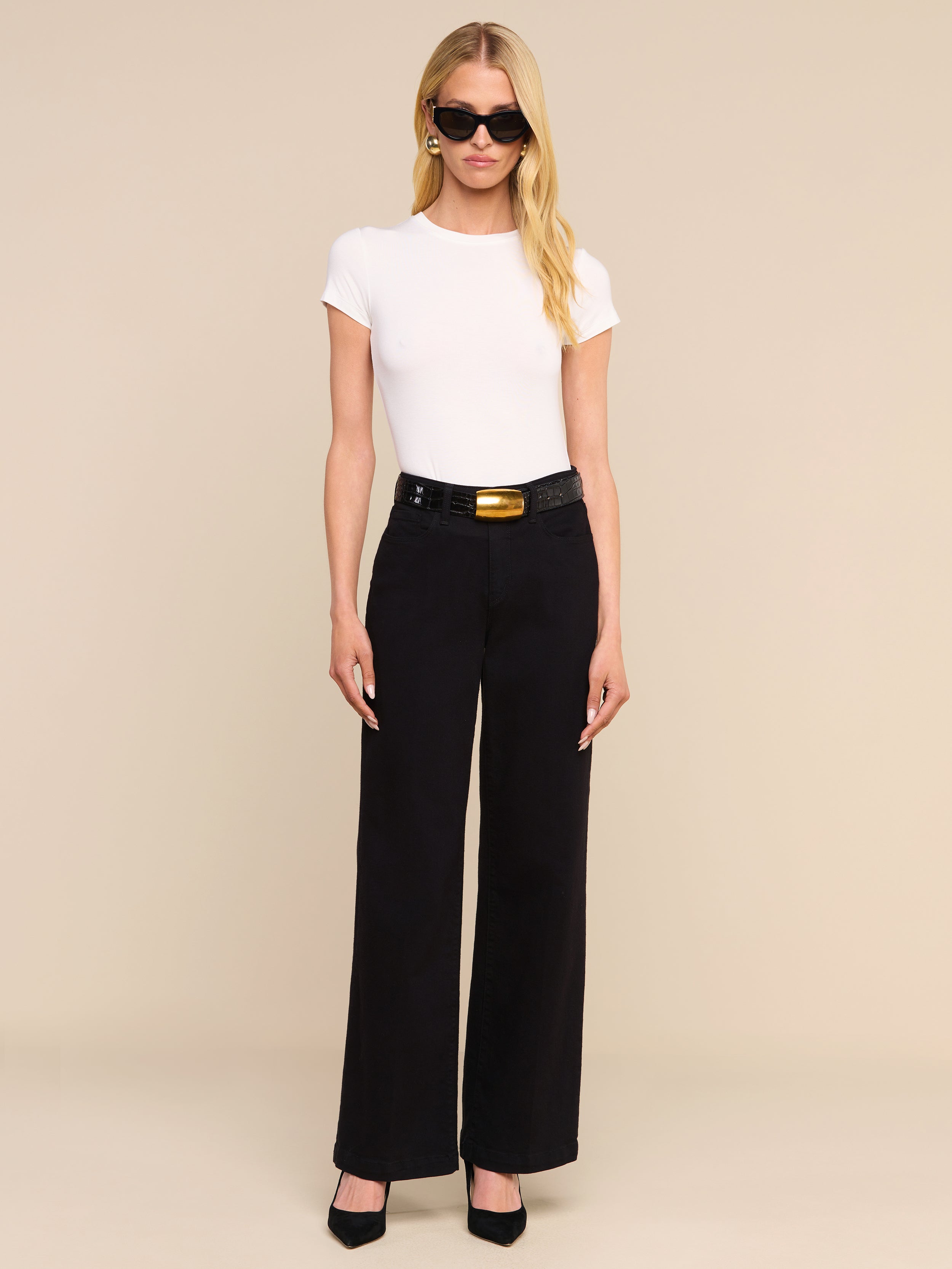 A woman with long blonde hair, wearing black sunglasses, a white short-sleeve top, and the Scottie Wide-Leg Jean paired with a black belt and black heels stands against a plain white background.