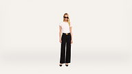 A woman with long blonde hair, wearing sunglasses, a white t-shirt, black high-waisted pants, and black heels, stands against a plain light background.