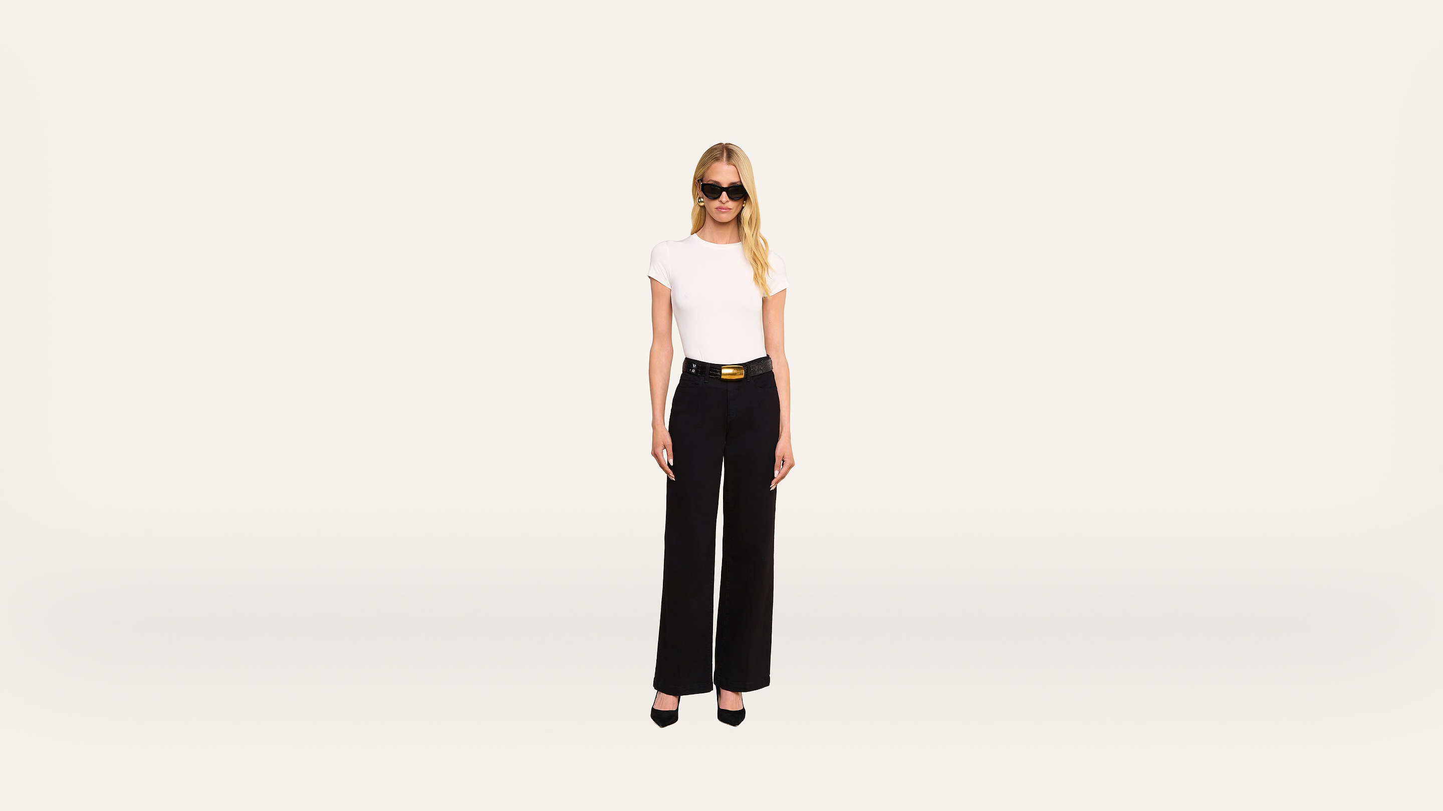 A woman with long blonde hair, wearing sunglasses, a white t-shirt, black high-waisted pants, and black heels, stands against a plain light background.