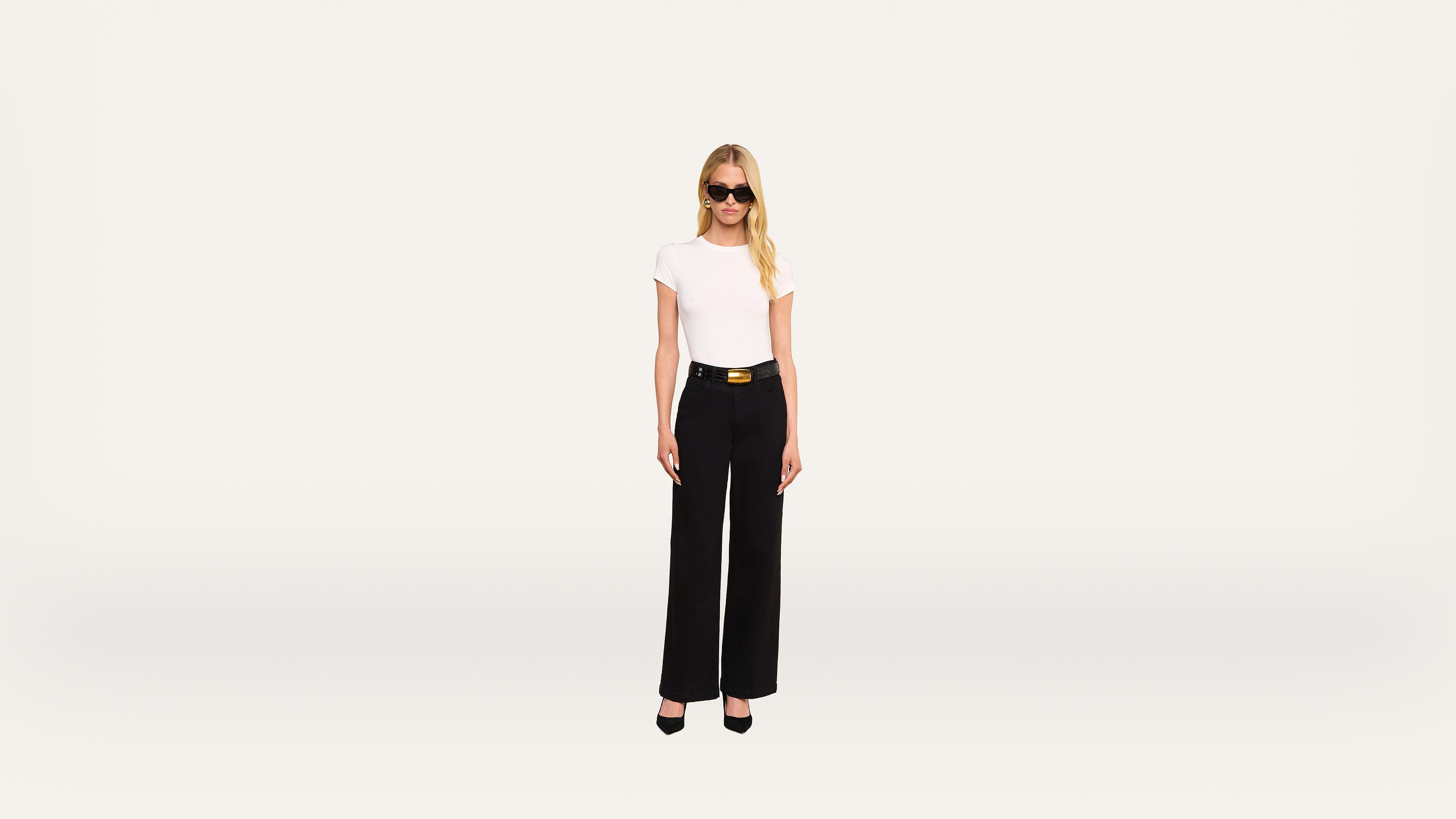 A woman with long blonde hair, wearing sunglasses, a white t-shirt, black high-waisted pants, and black heels, stands against a plain light background.