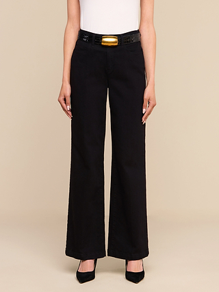 Una persona indossa i jeans Scottie Wide-Leg, un modello a vita alta nero realizzato in denim elasticizzato di alta qualità, abbinati a una cintura nera con fibbia dorata, un top bianco senza maniche e tacchi neri a punta, in piedi davanti a uno sfondo beige.