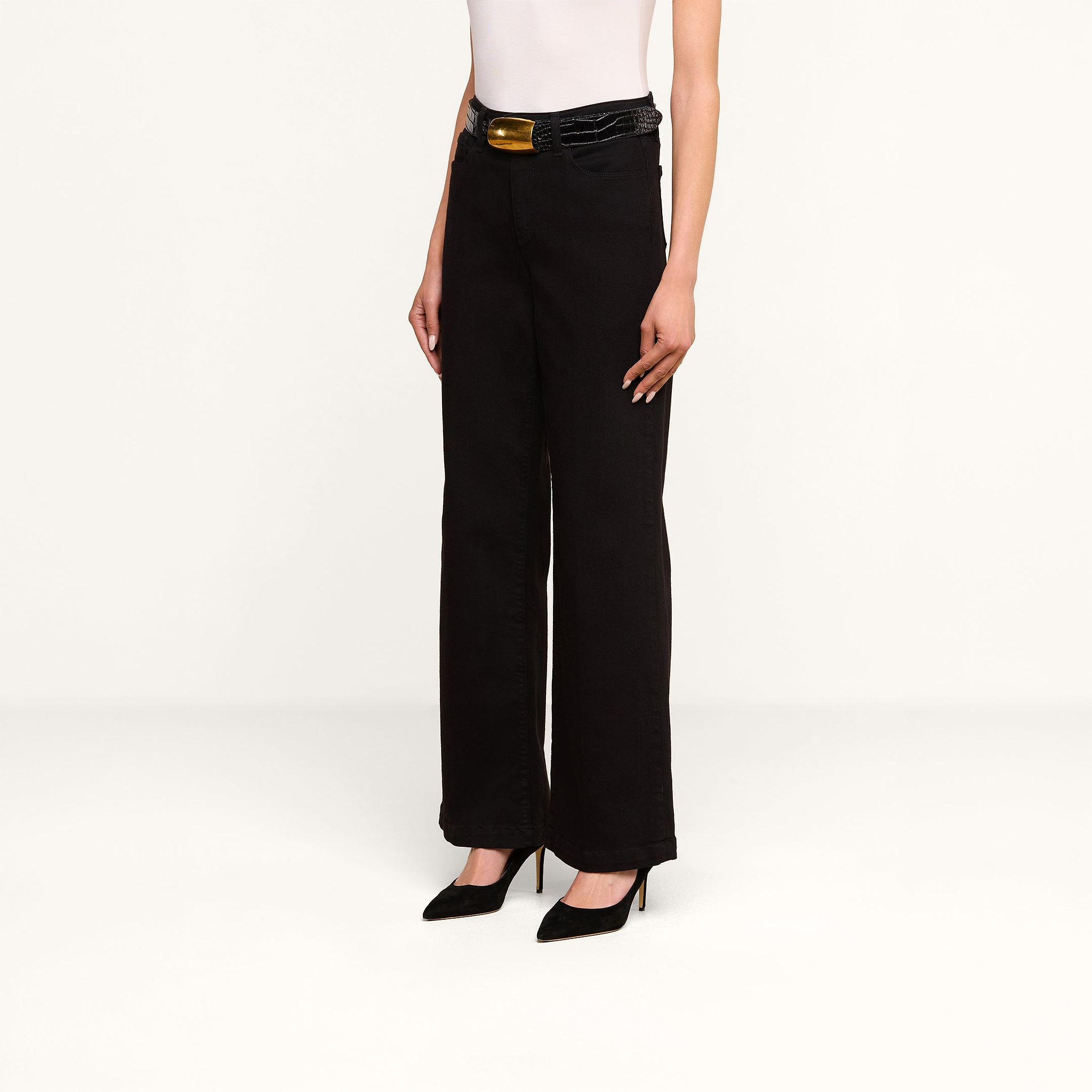 A person stands against a plain white background wearing the Scottie Wide-Leg Jean in black high-rise premium stretch denim, paired with a white sleeveless top, black high heels, and a black belt with a gold buckle.