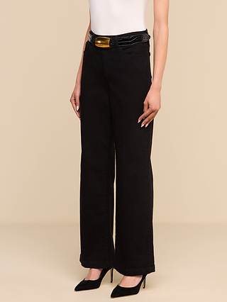 Una persona que lleva una camiseta blanca sin mangas, unos vaqueros Scottie Wide-Leg confeccionados en denim elástico de primera calidad, un cinturón negro con una gran hebilla dorada y unos zapatos de tacón negros posa frente a un fondo liso de color beige.