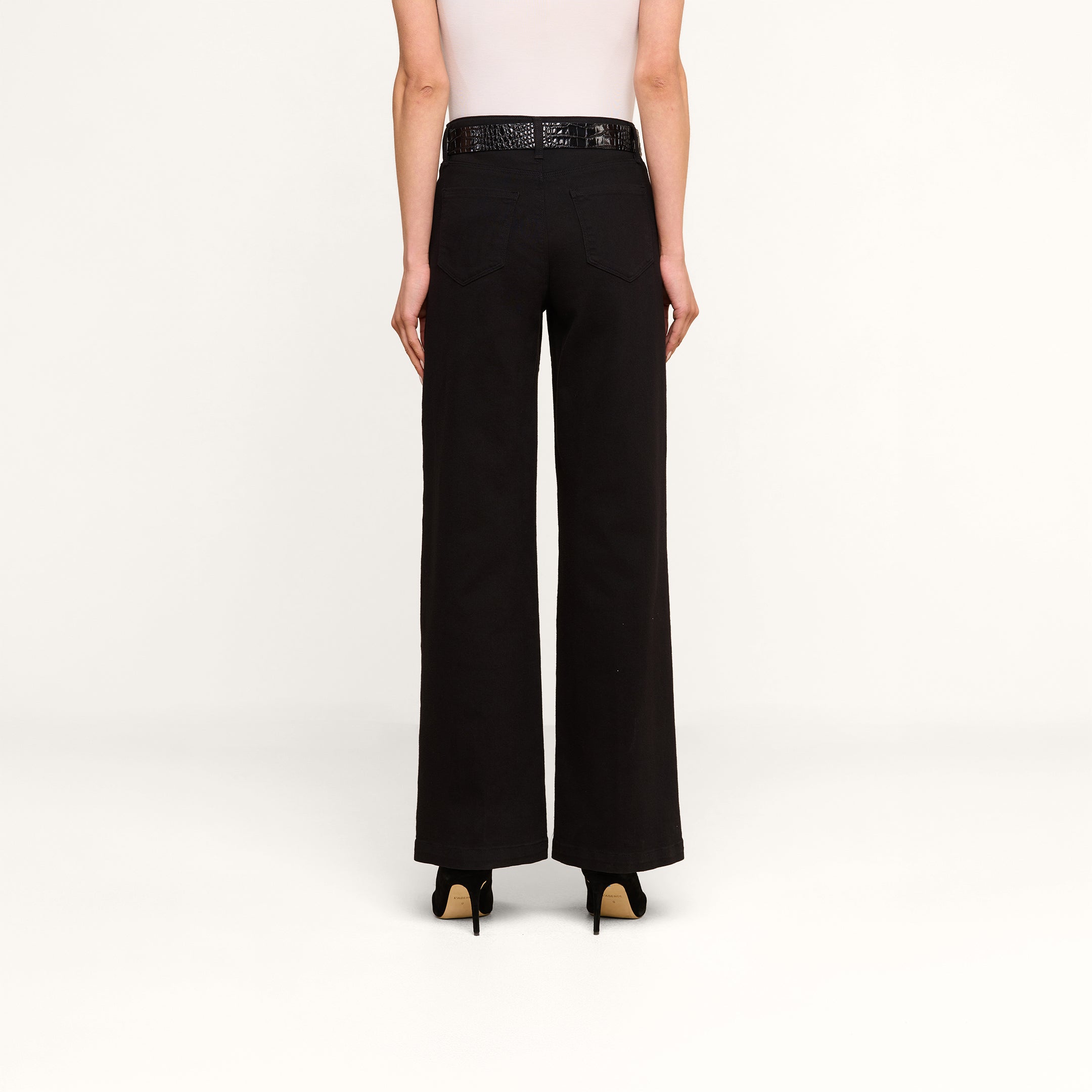 A person stands facing away on a plain white background, wearing a white sleeveless top, the Scottie Wide-Leg Jean in black high-rise, a black belt, and black heels.