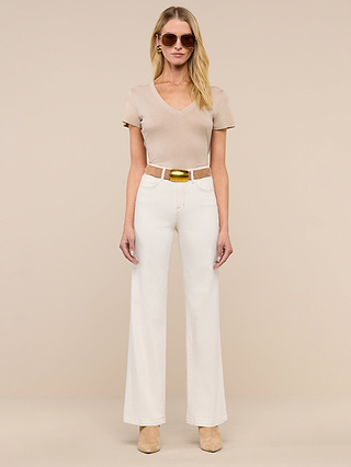 A woman stands against a beige background, wearing sunglasses, a beige short-sleeve top, the Scottie Wide-Leg Jean in white with a white belt featuring a gold buckle, and beige shoes.