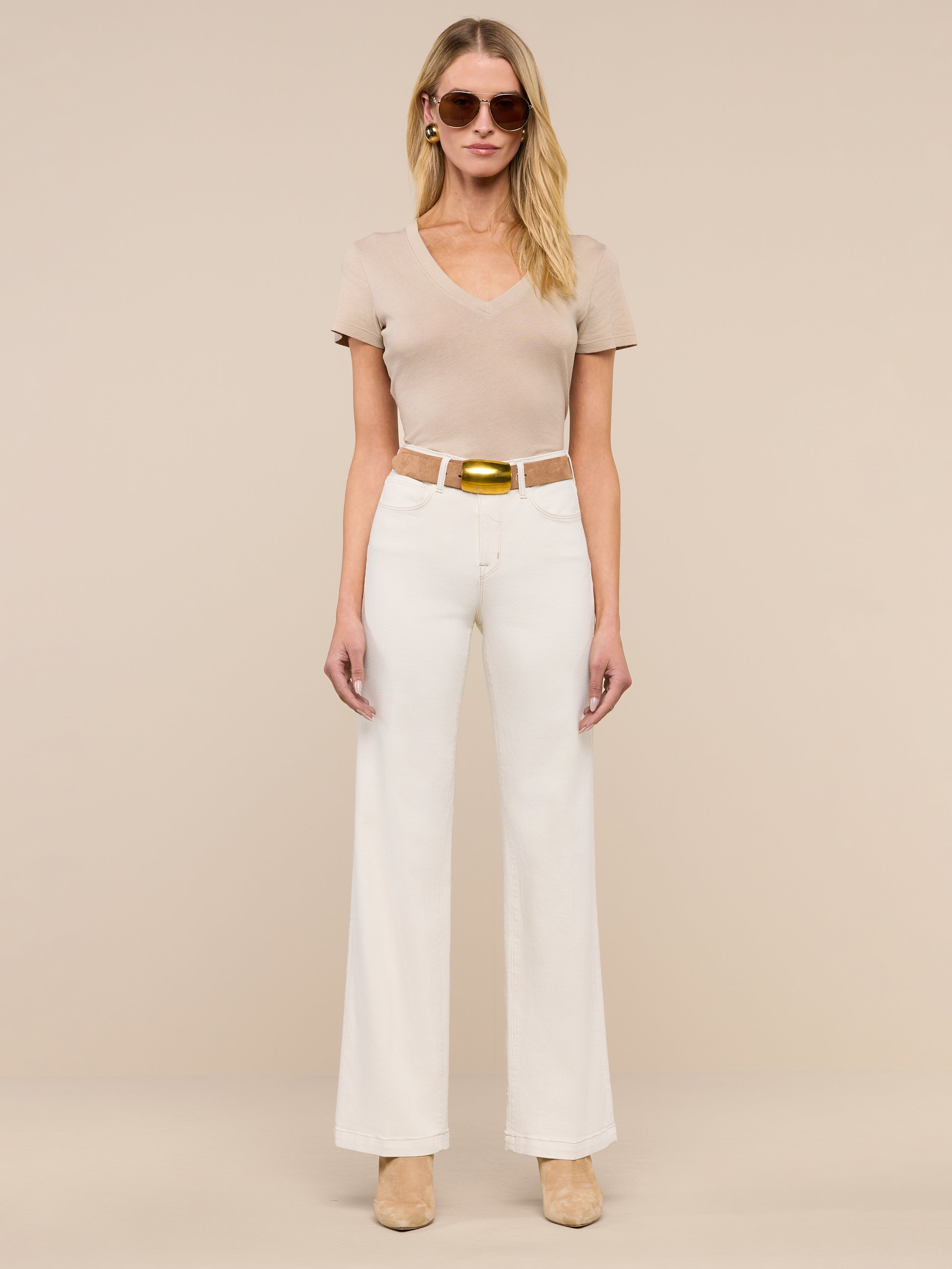 A woman wearing beige sunglasses, a short-sleeve beige top, the Scottie Wide-Leg Jean in cream with a tan belt, and beige heels stands against a plain light background.