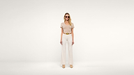 Woman standing against a plain white background, wearing sunglasses, a beige short-sleeve top, wide-legged white pants, a tan belt, and beige shoes. She is facing forward with her arms relaxed by her sides.