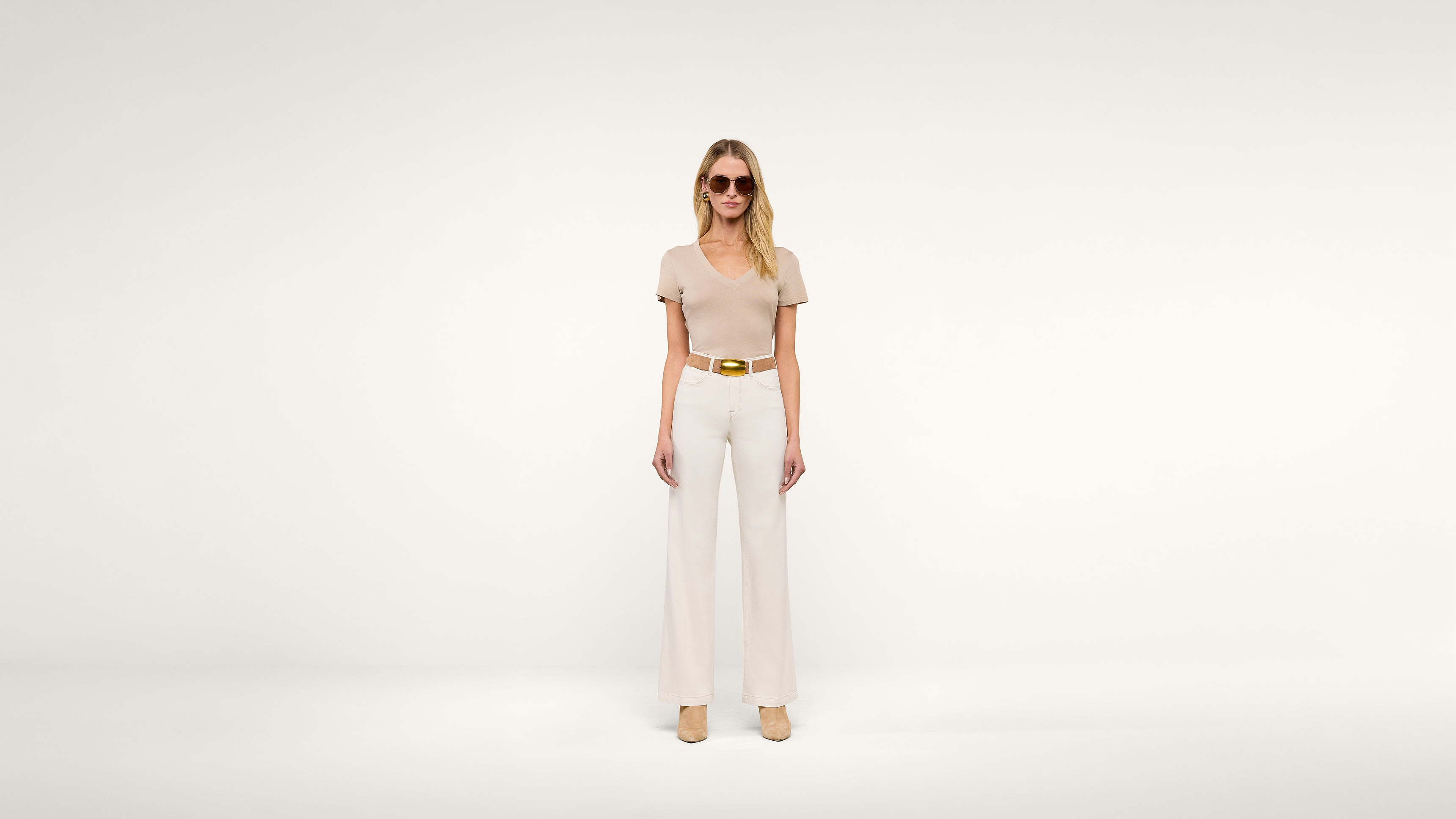 Woman standing against a plain white background, wearing sunglasses, a beige short-sleeve top, wide-legged white pants, a tan belt, and beige shoes. She is facing forward with her arms relaxed by her sides.
