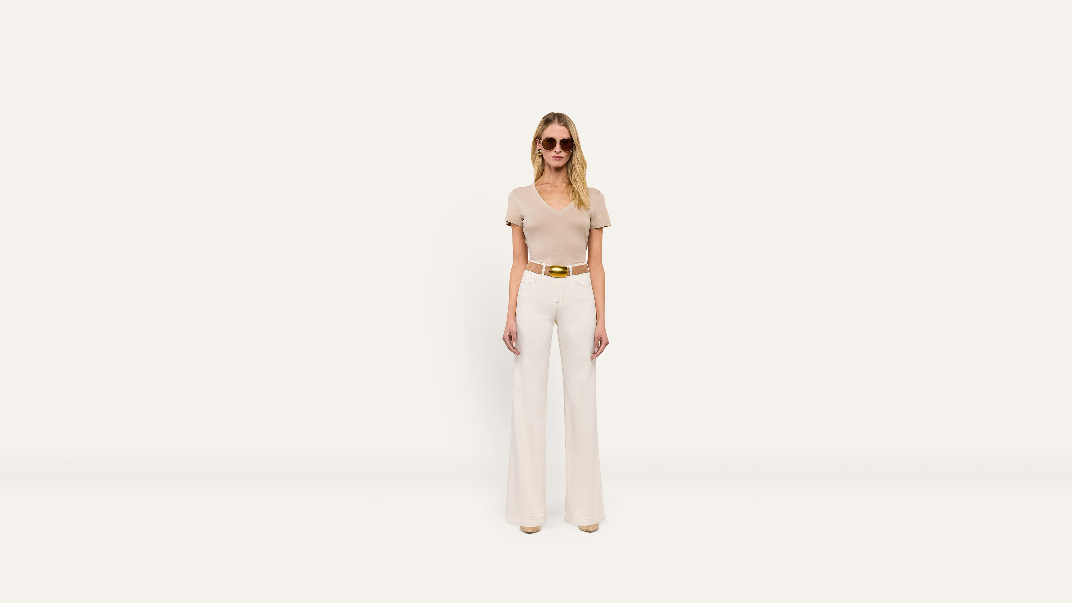 A woman wears a tan v-neck t-shirt, white wide-leg trousers with a gold-buckle belt, and brown sunglasses.