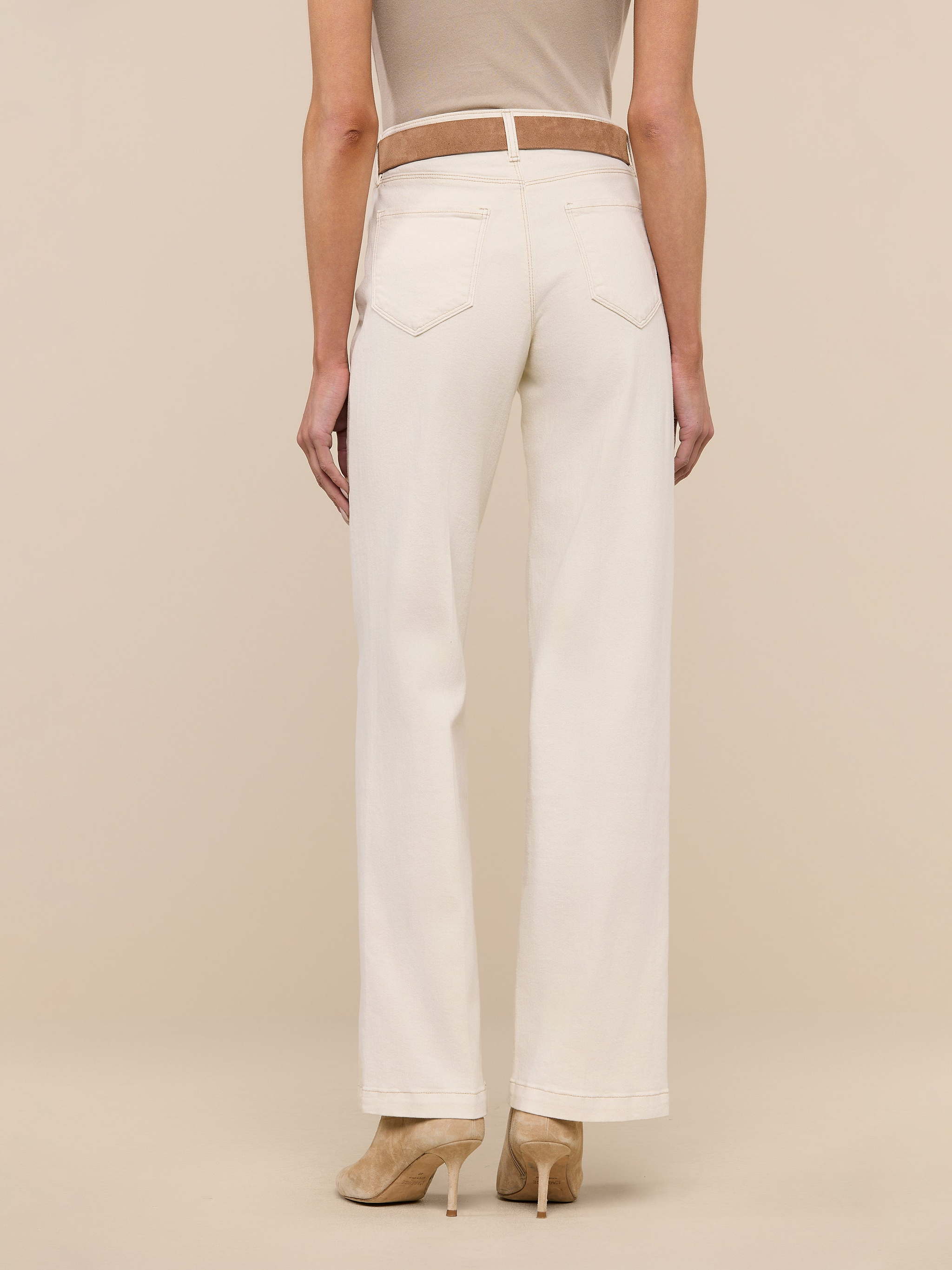 A woman stands facing away against a plain white background, wearing the Scottie Wide-Leg Jean in a cream color, paired with a beige sleeveless top and beige heeled shoes.