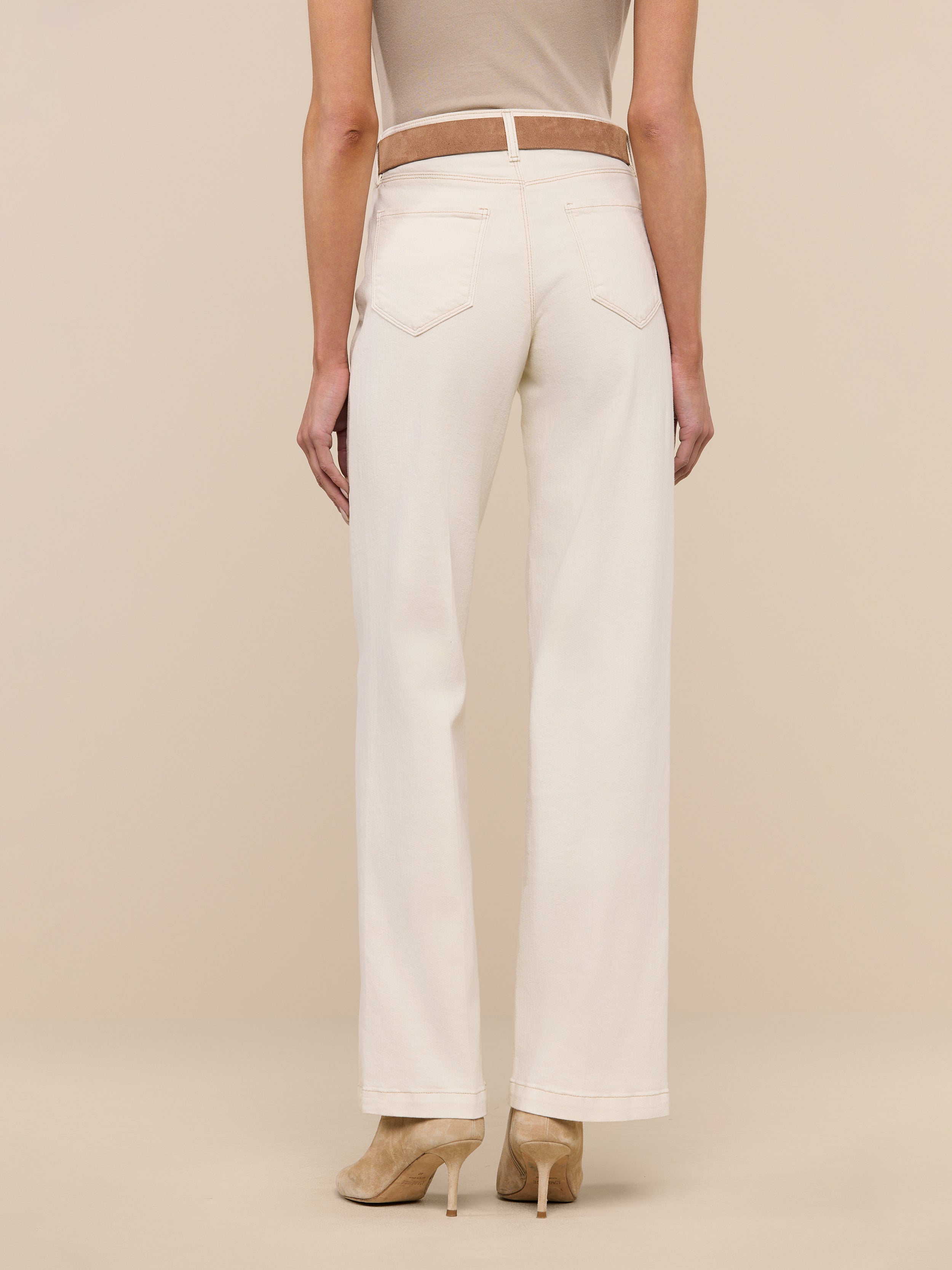 A woman stands facing away against a plain white background, wearing the Scottie Wide-Leg Jean in a cream color, paired with a beige sleeveless top and beige heeled shoes.