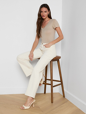 A woman with long brown hair sits on a wooden stool in a minimal, light room, wearing a beige short-sleeve top, Scottie Wide-Leg Jean with a high-rise waistband, and cream heeled sandals on wooden flooring.
