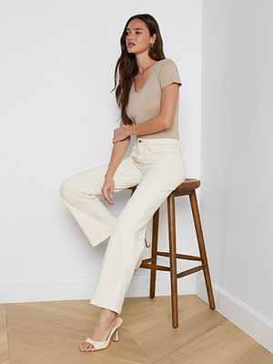 A woman with long brown hair sits on a wooden stool against a white wall, wearing a beige short-sleeve top, the Scottie Wide-Leg Jean in cream with a high-rise waistband, and beige heeled sandals. She gazes to the side with a relaxed expression.