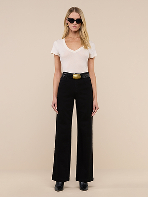 A woman with long blonde hair wears black sunglasses, a white short-sleeve V-neck top, Scottie Wide-Leg Jean with a belt, and black shoes, standing against a plain beige background.